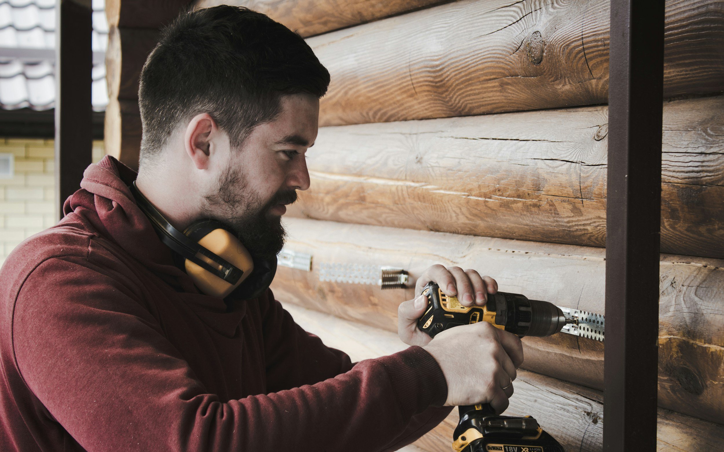 A man with dark hair and a beard using a yellow and black cordless drill to work on a wooden wall in a workshop or home improvement setting.