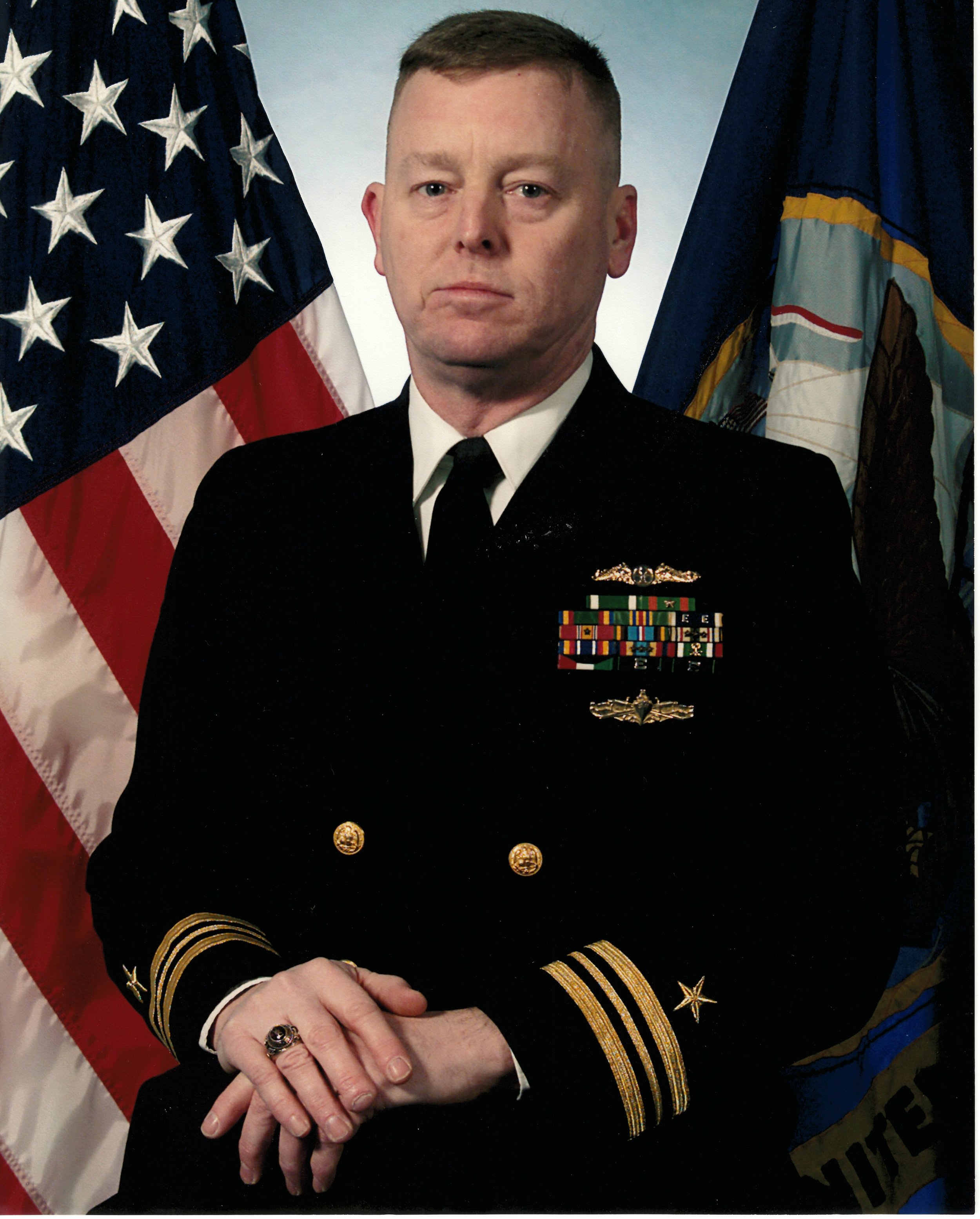 Mike France in his U.S. Navy uniform with gold and black embellishments, standing in front of American and military flags.