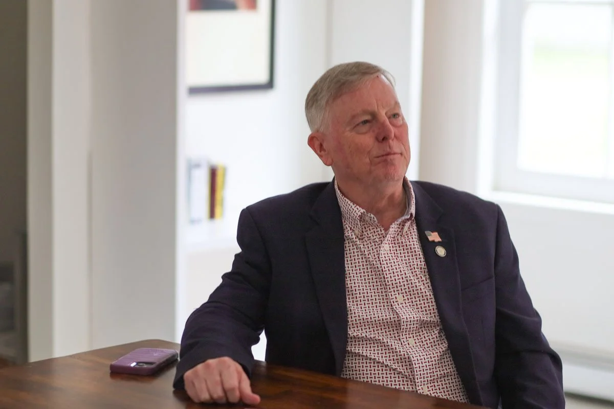 Mike France for State Senate sitting at a wooden table in a bright room with white walls, wearing a dark blazer, a patterned shirt, and a small American flag pin.