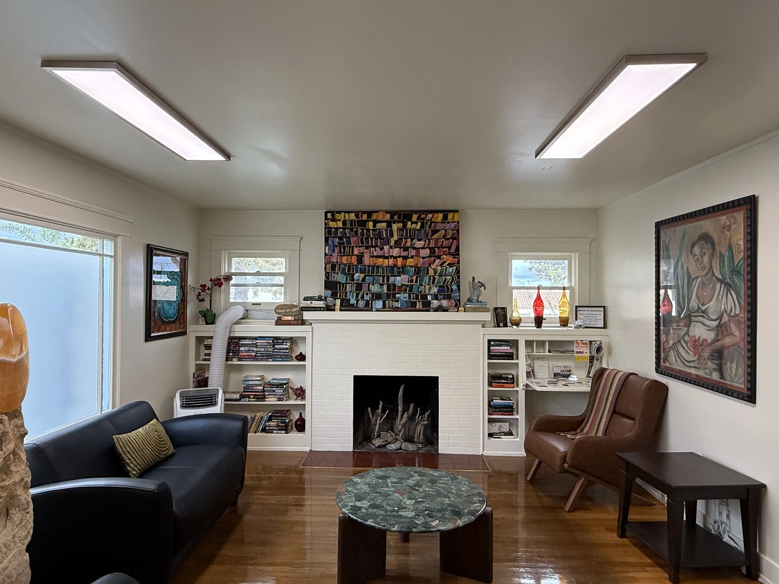 Teresa Monzon Dental Looby with white brick fireplace, bookshelves, artwork, and seating including a black sofa with yellow pillows and a brown leather armchair, illuminated by natural light from windows.