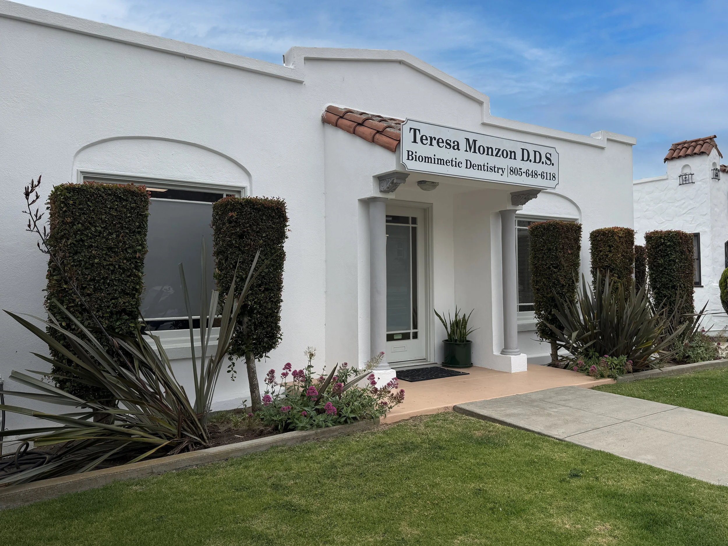 White building with a sign for Teresa Monzon D.D.S. Biomimetic Dentistry, surrounded by identical bushes and plants, under a blue sky.