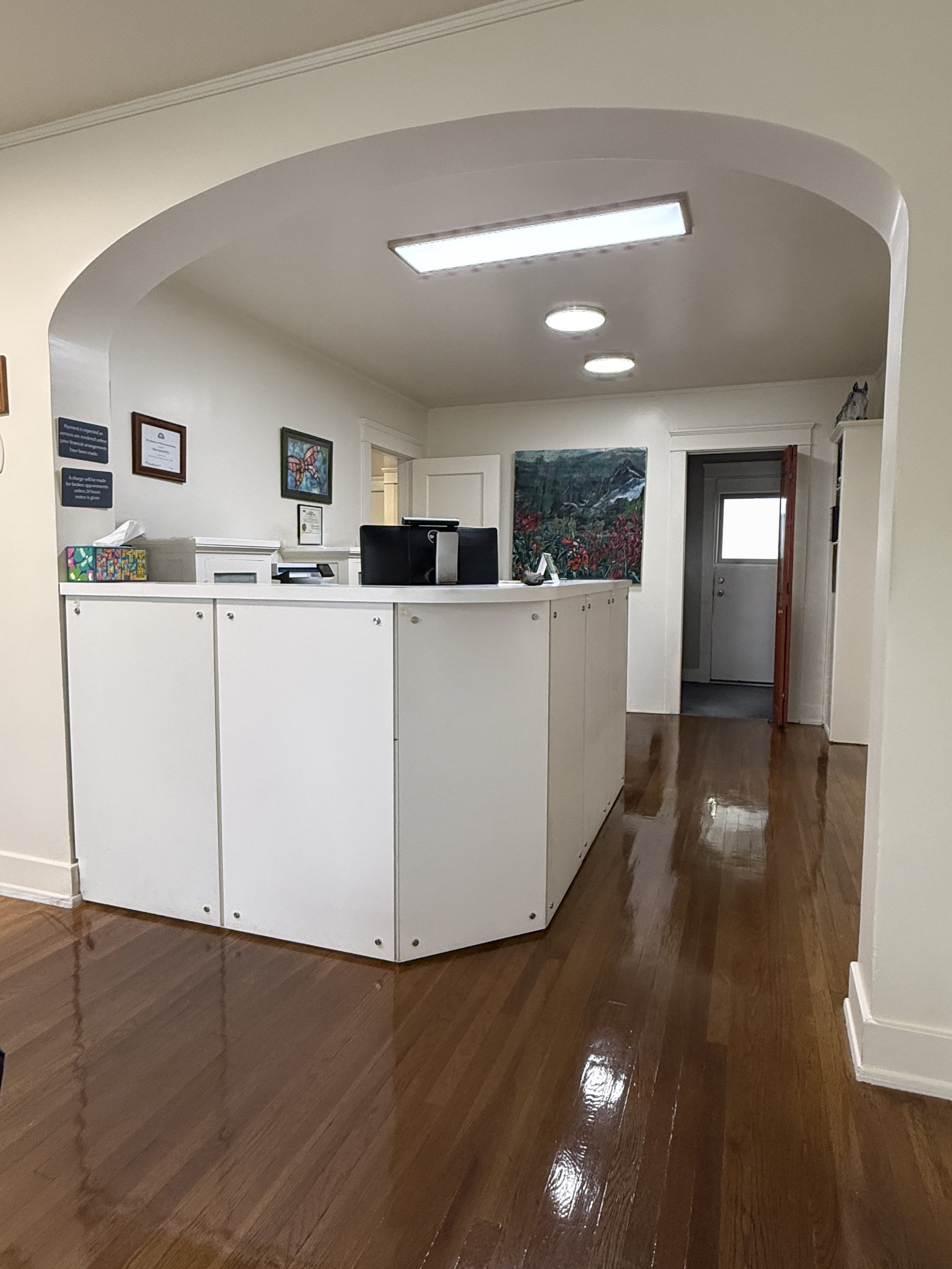 A reception area with a white counter, computer, framed artwork, and paintings on the wall. Wooden floors and a door in the background.