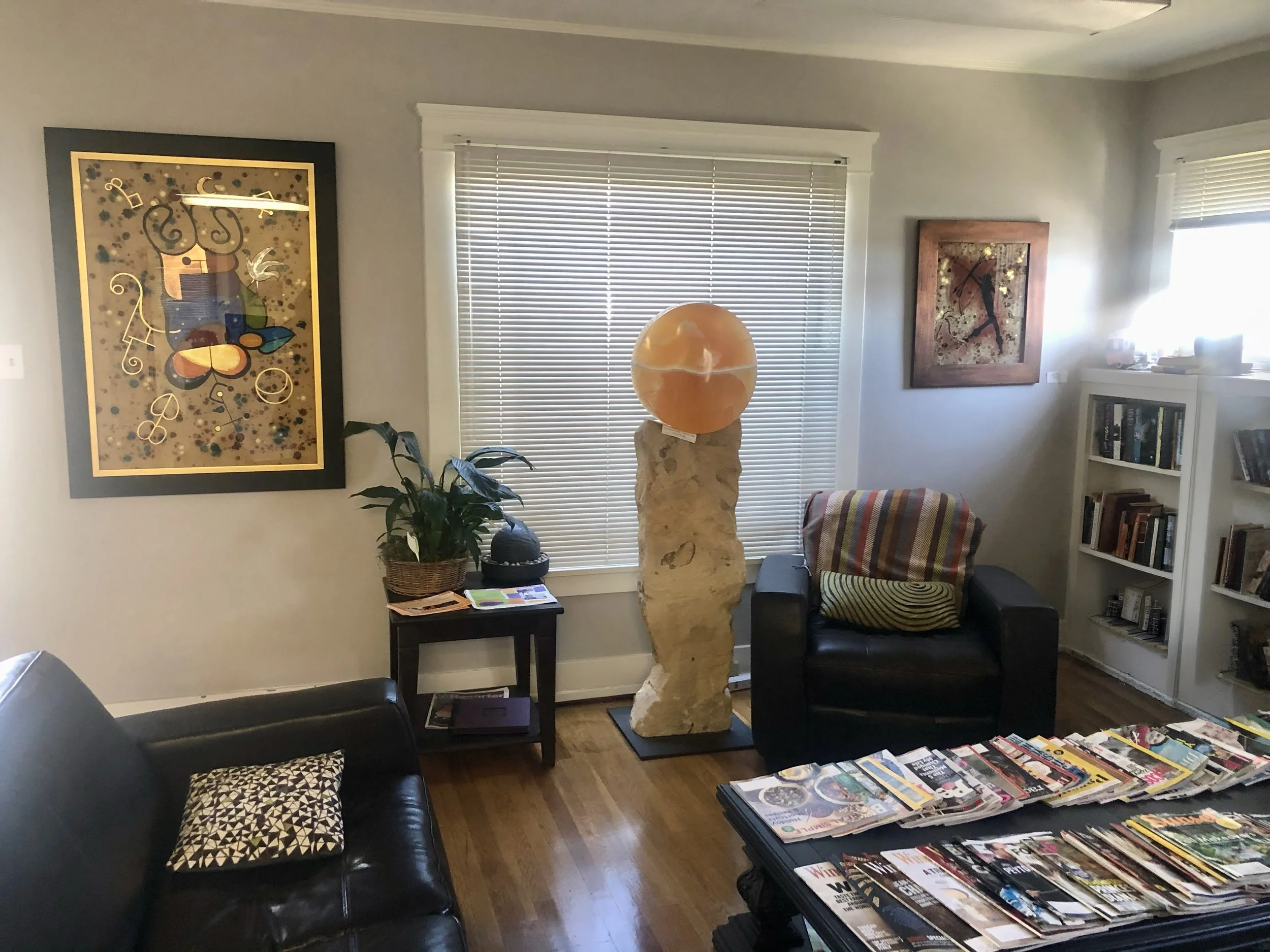 Lobby with large windows with blinds, artwork on the walls, a black leather couch with a patterned pillow, a small side table with magazines, a bookshelf, and a marble sculpture with an orange sphere on top.