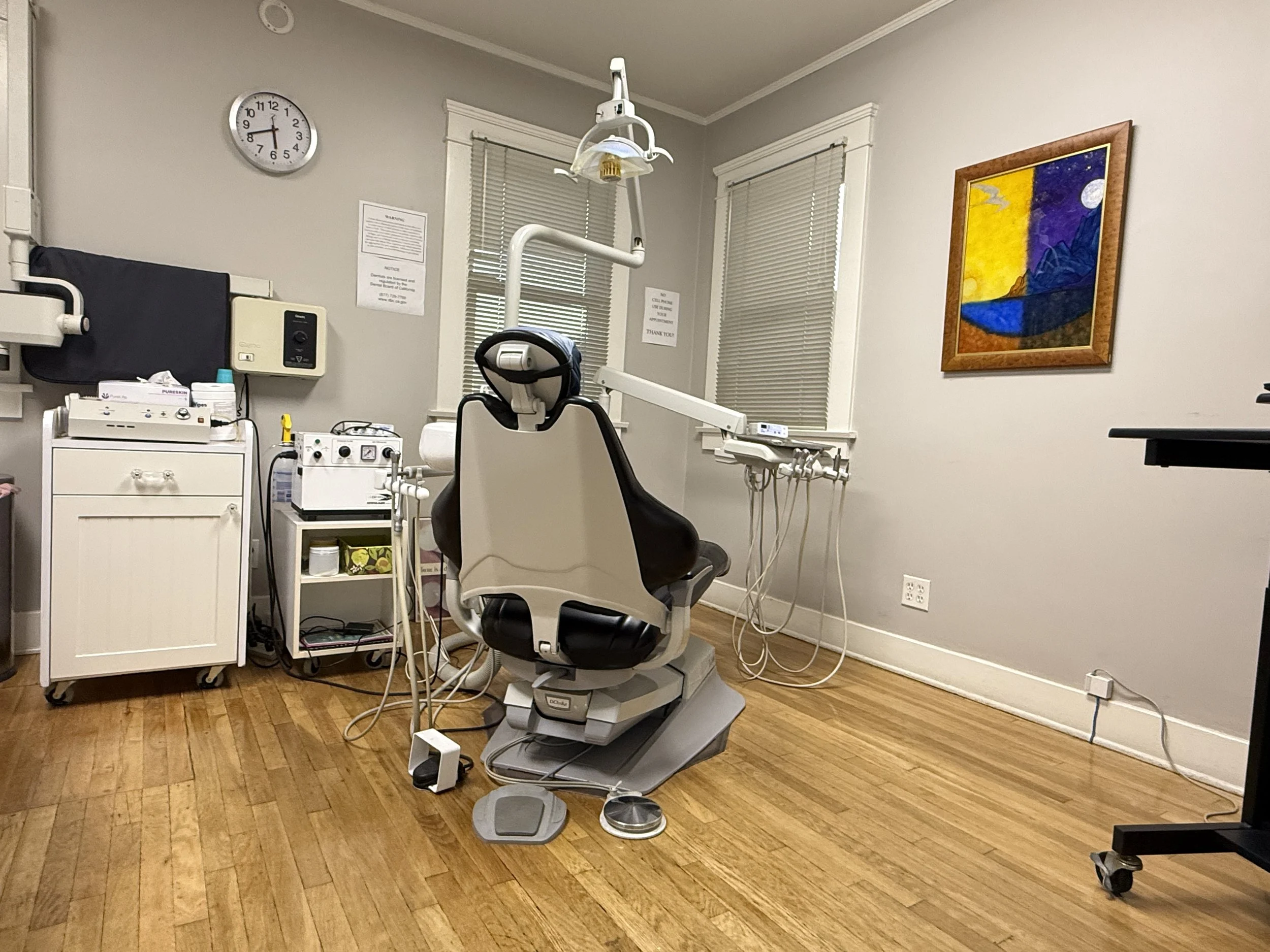 A dental examination room with a dental chair at the center, surrounded by medical equipment, and artwork on the walls.