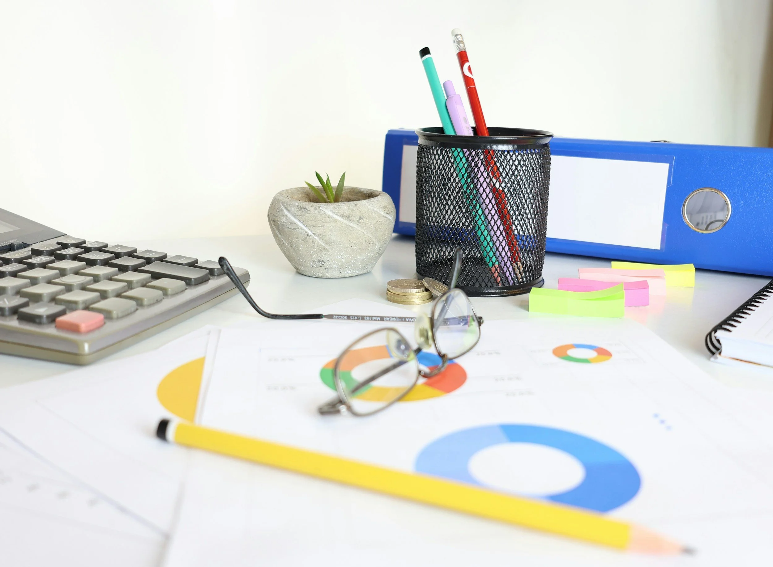 Office desk with a black pen holder containing three pens, a small potted succulent plant, a blue binder, a pair of glasses, a calculator, some colorful sticky notes, a coin, and a paper with a pie chart.