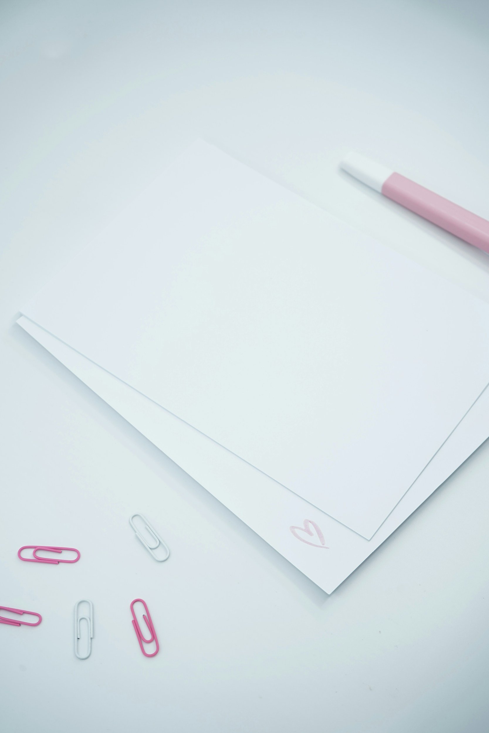 Flat lay of a white desk with scattered pink and white paper clips, a white sheet of paper with a pink heart drawing, and an eraser with a pink sleeve.