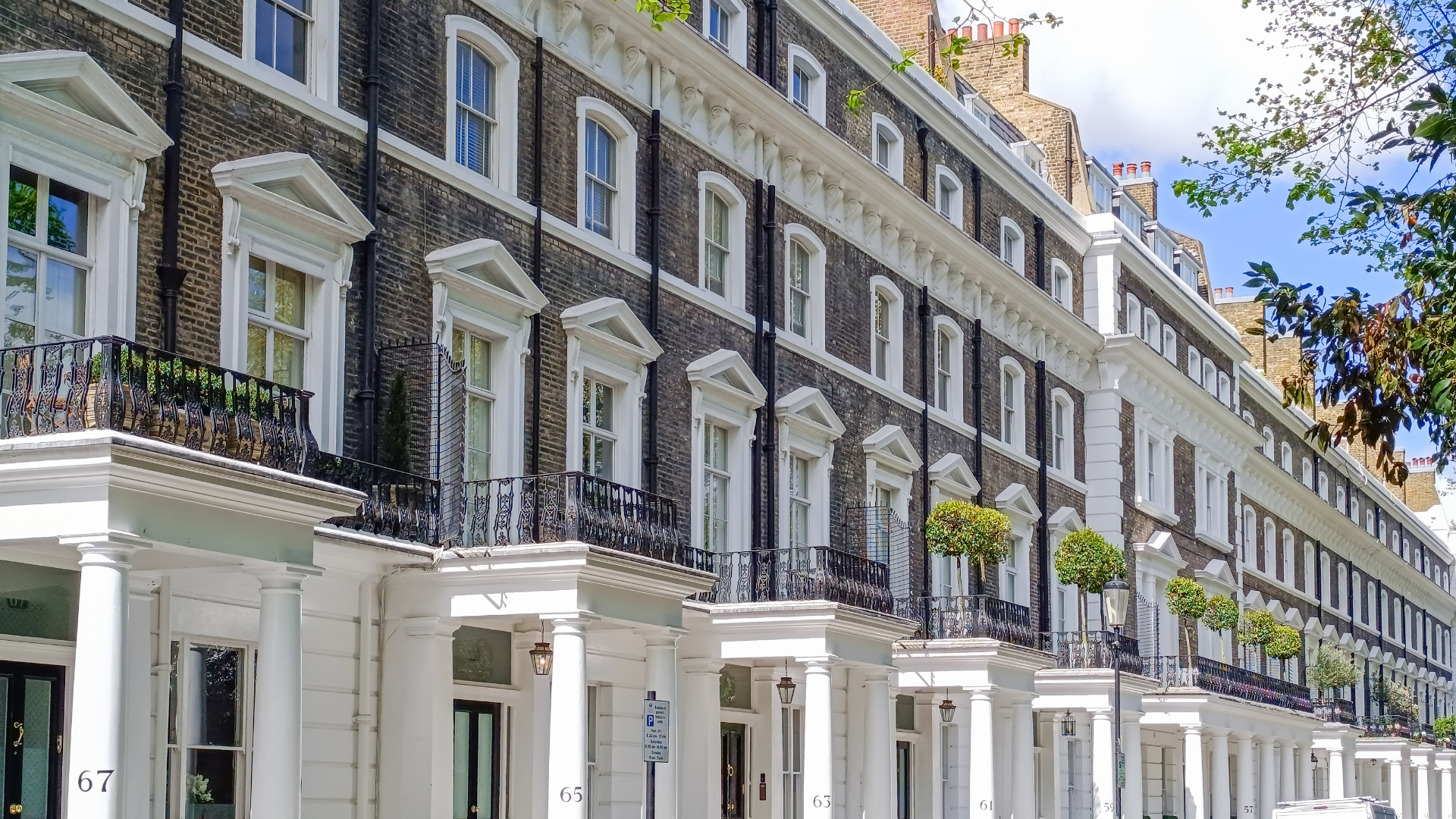 Rows of classic European-style townhouse buildings with white and brick facades, black wrought iron balconies, and tall windows, lined along a street with small trees and street lamps.