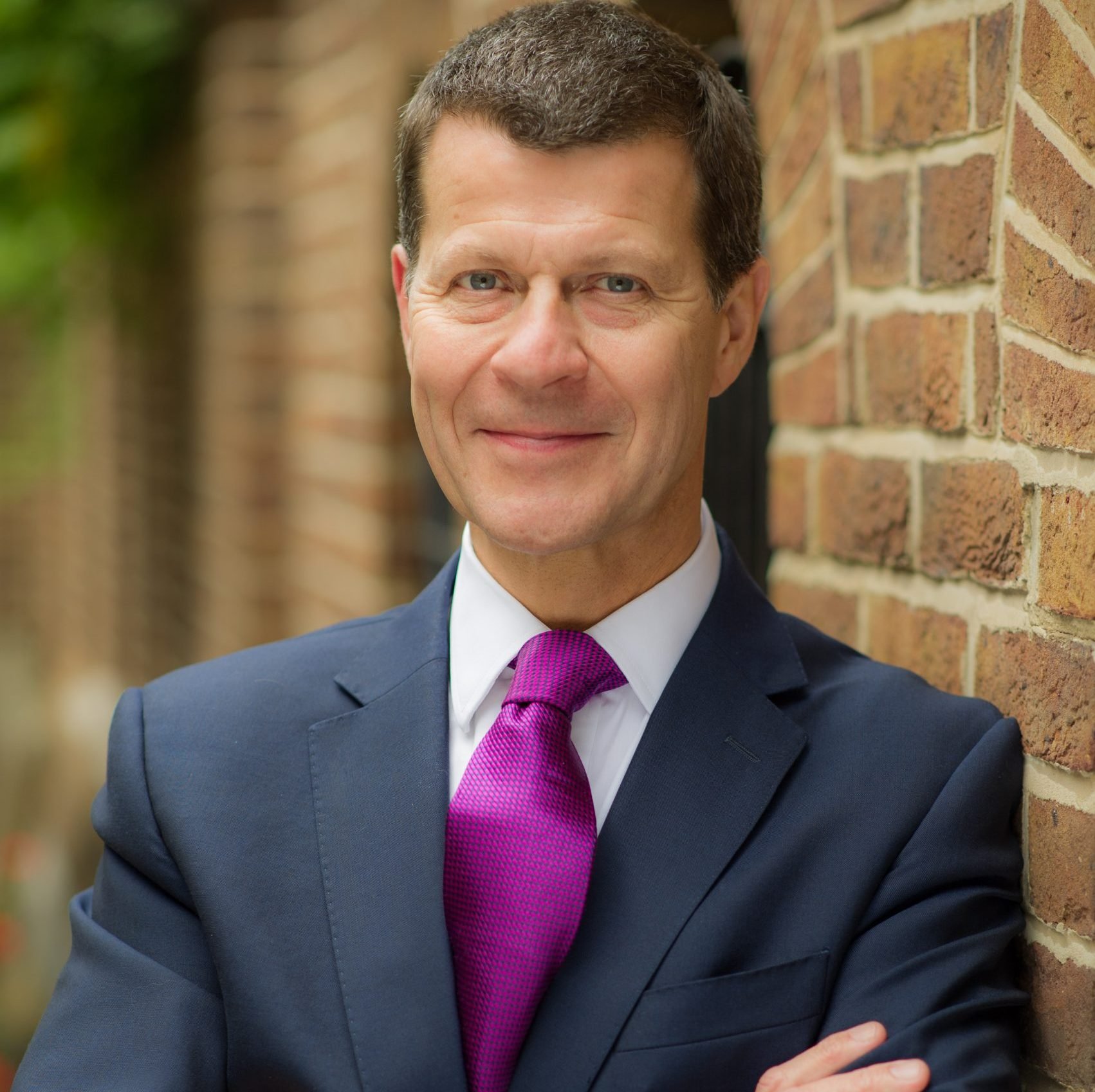 A portrait of Tony Horrell,  in a dark suit with a white shirt and a purple tie, standing outdoors against a brick wall, smiling with arms crossed.