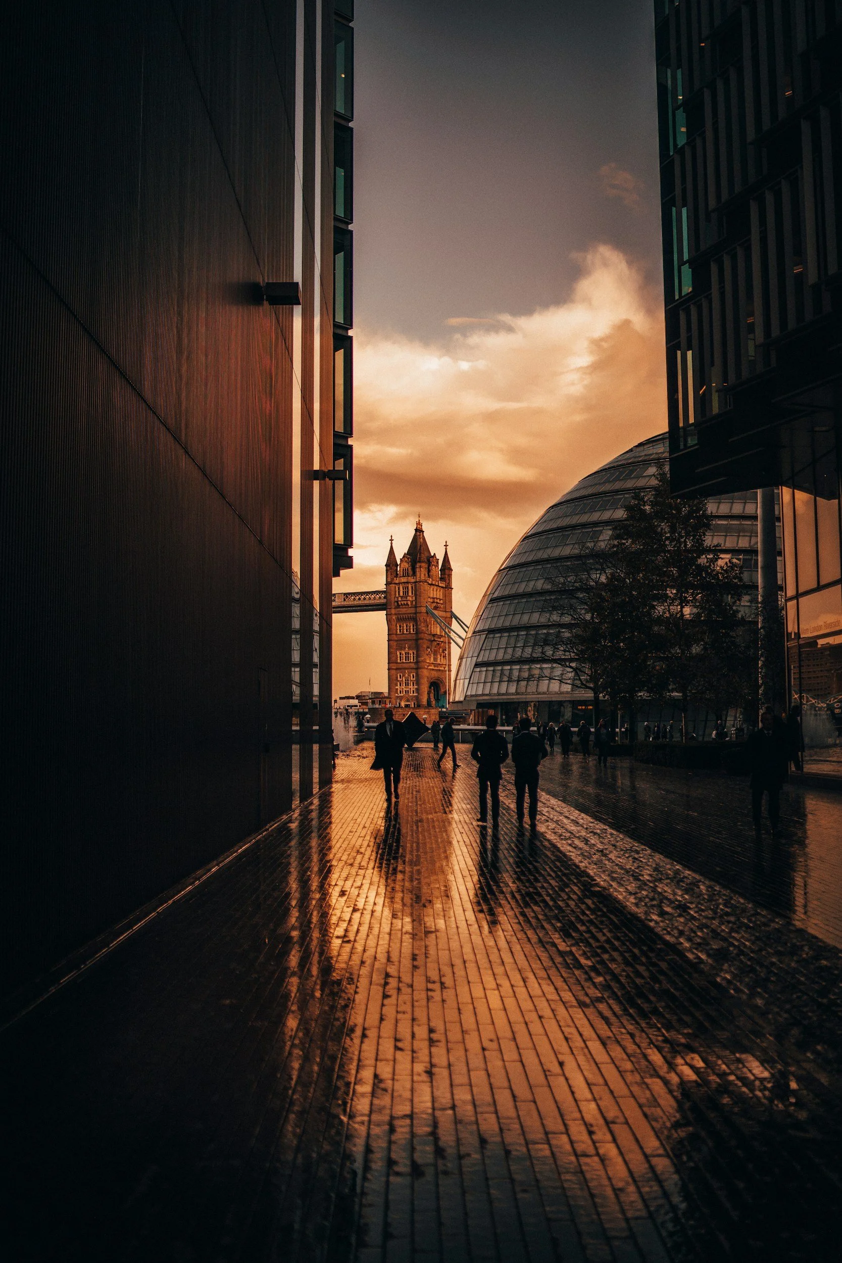 People walking on a wet brick sidewalk near modern buildings with glass windows at sunset, with Tower Bridge visible in the background.