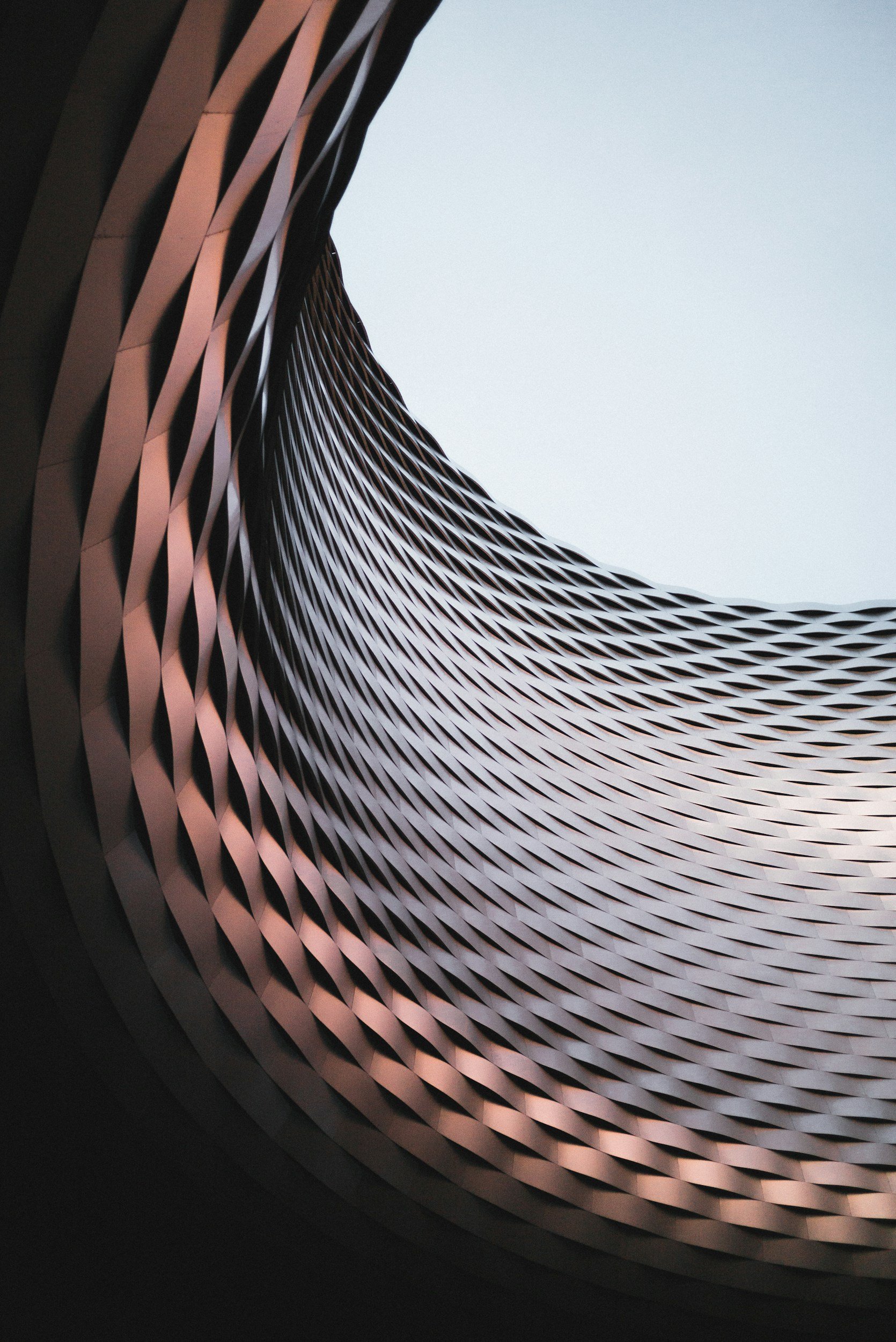 An abstract architectural structure with a spiral pattern of pinkish-beige geometric panels, viewed from underneath against a clear sky.