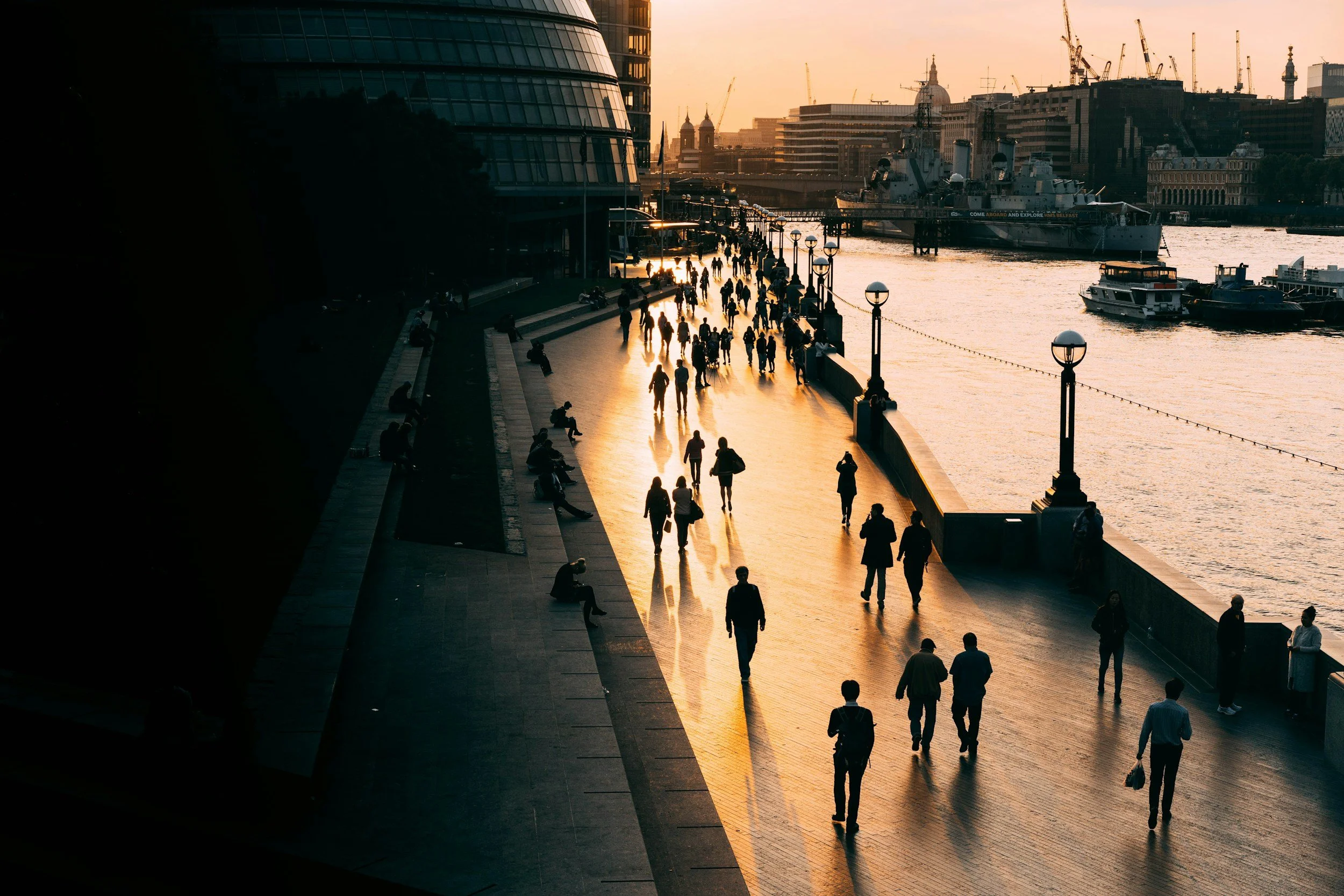 People walking along a riverside promenade in London during sunset, with boats docked in the water and city buildings in the background.