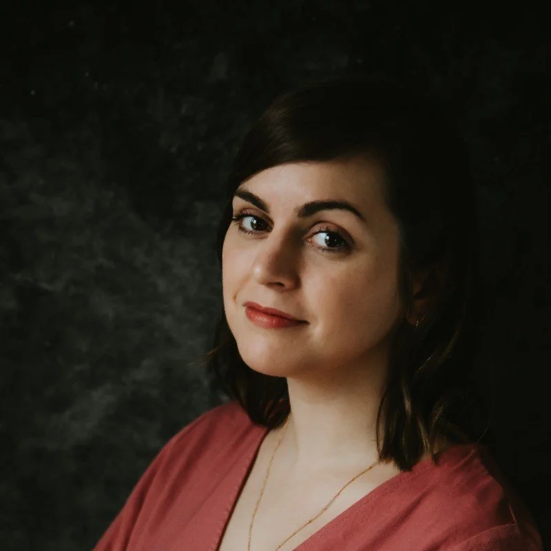A portrait of Helen Herbert - a woman with short dark hair, wearing a pink top, standing against a dark textured background, looking at the camera with a slight smile.