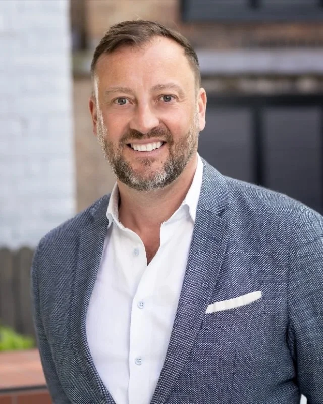 Portrait of a Kristen Zeilerbauer - a smiling man with short hair and a beard, wearing a light blue blazer and white shirt, standing outdoors in front of a brick building.