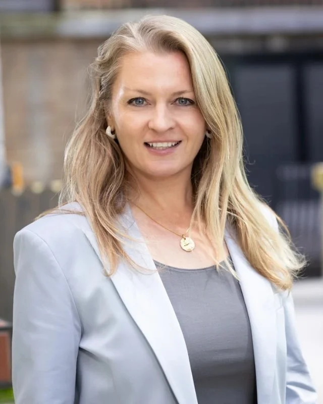 A portrait of Nina Zeilerbauer - a woman with blonde hair, wearing a light blazer over a grey top, smiling outdoors.