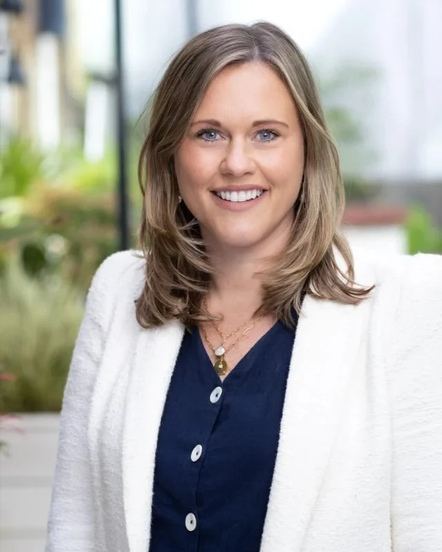 A portrait of Clare Coe - a woman with shoulder-length light brown hair, blue eyes, and a warm smile, wearing a white blazer over a navy blue blouse, sitting in a bright indoor space with greenery in the background.
