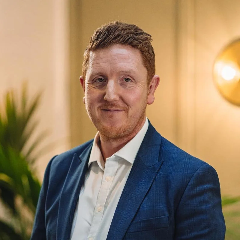 A man with reddish hair and a beard wearing a white shirt and a blue blazer, standing indoors with a blurred background featuring plants and warm lighting.