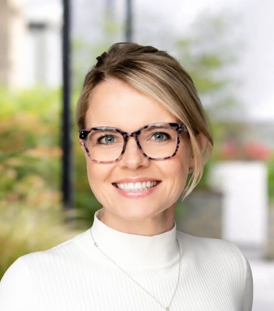 A portrait of Rachel Maguire - smiling woman with short blonde hair, wearing glasses and a white turtleneck, indoors with a blurred window and greenery in the background.