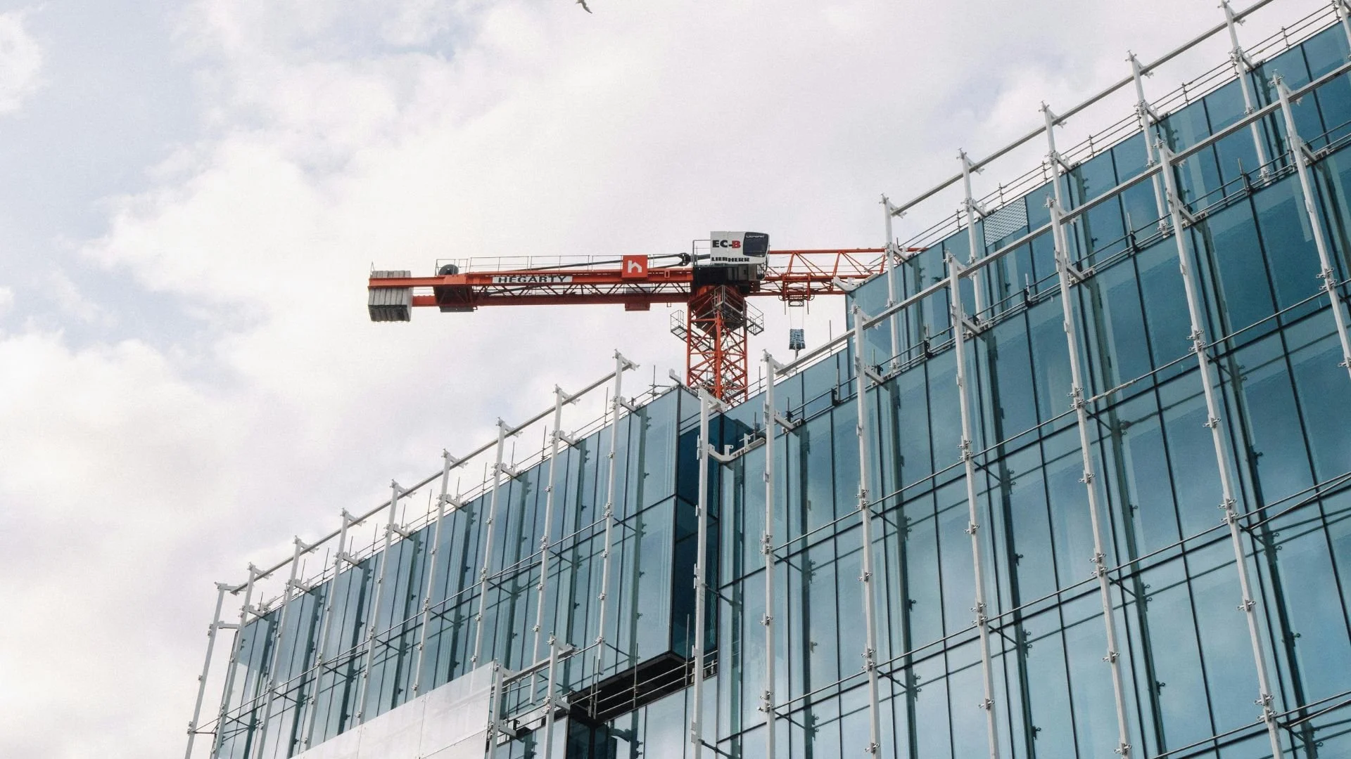 A construction crane on top of a modern glass building under construction with scaffolding on the facade.