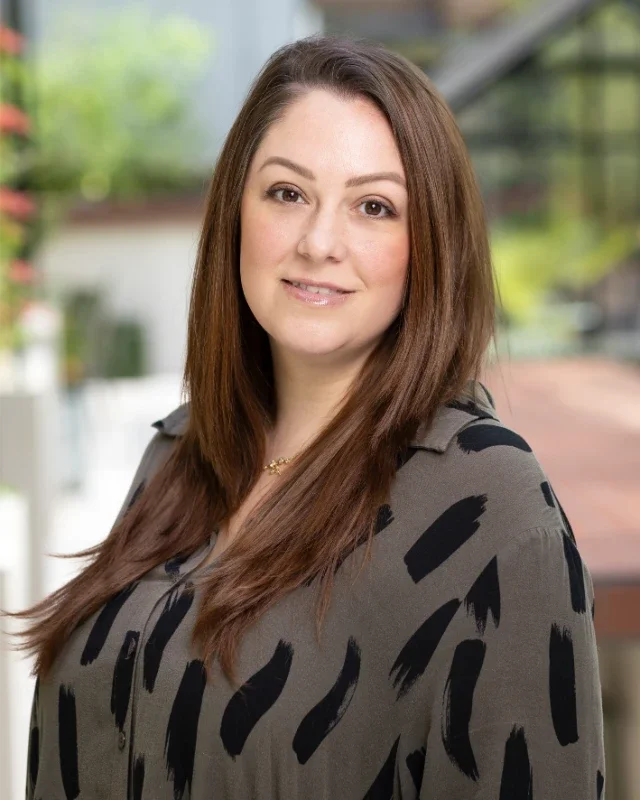 A portrait of Gemma Northeast - a woman with long brown hair smiling outdoors, wearing a grey shirt with black abstract patterns.
