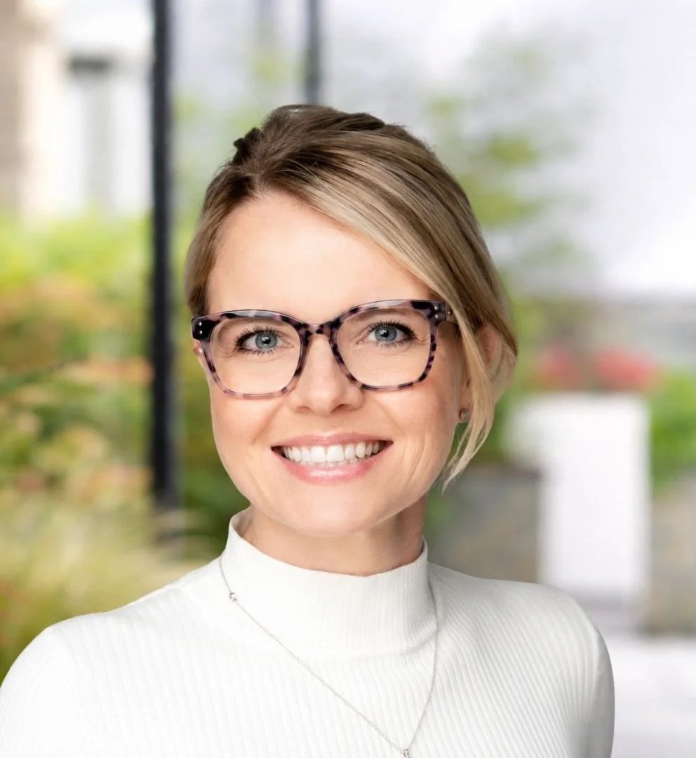 Rachel Maguire - a smiling woman with glasses, blonde hair, and a white turtleneck standing outdoors with a blurred background of greenery and buildings.