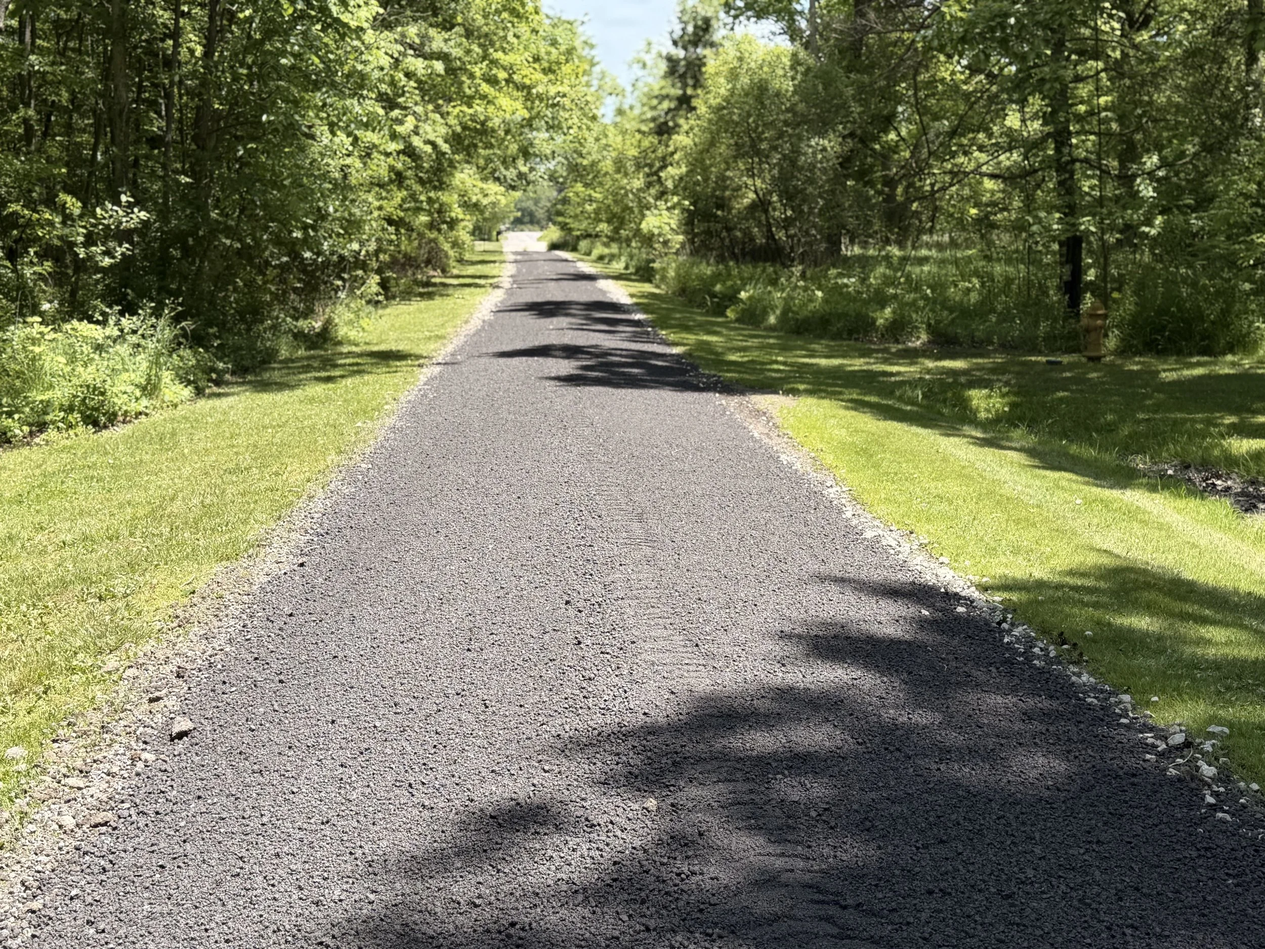 A gravel country road surrounded by green trees on both sides, with blue skies overhead.
