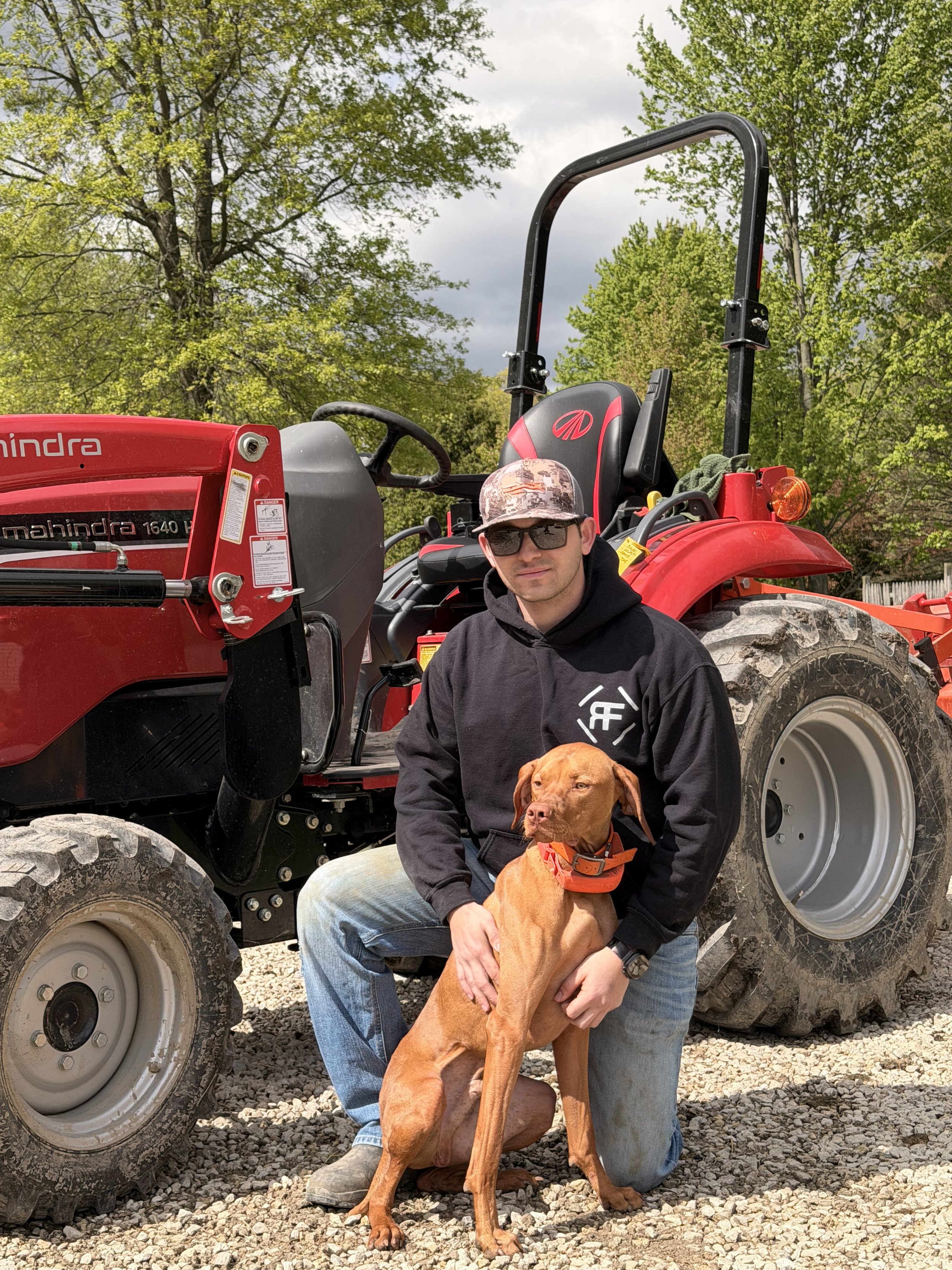 A man in sunglasses and a black hoodie kneeling next to a brown dog with a red collar, in front of a red tractor with a green background of trees.