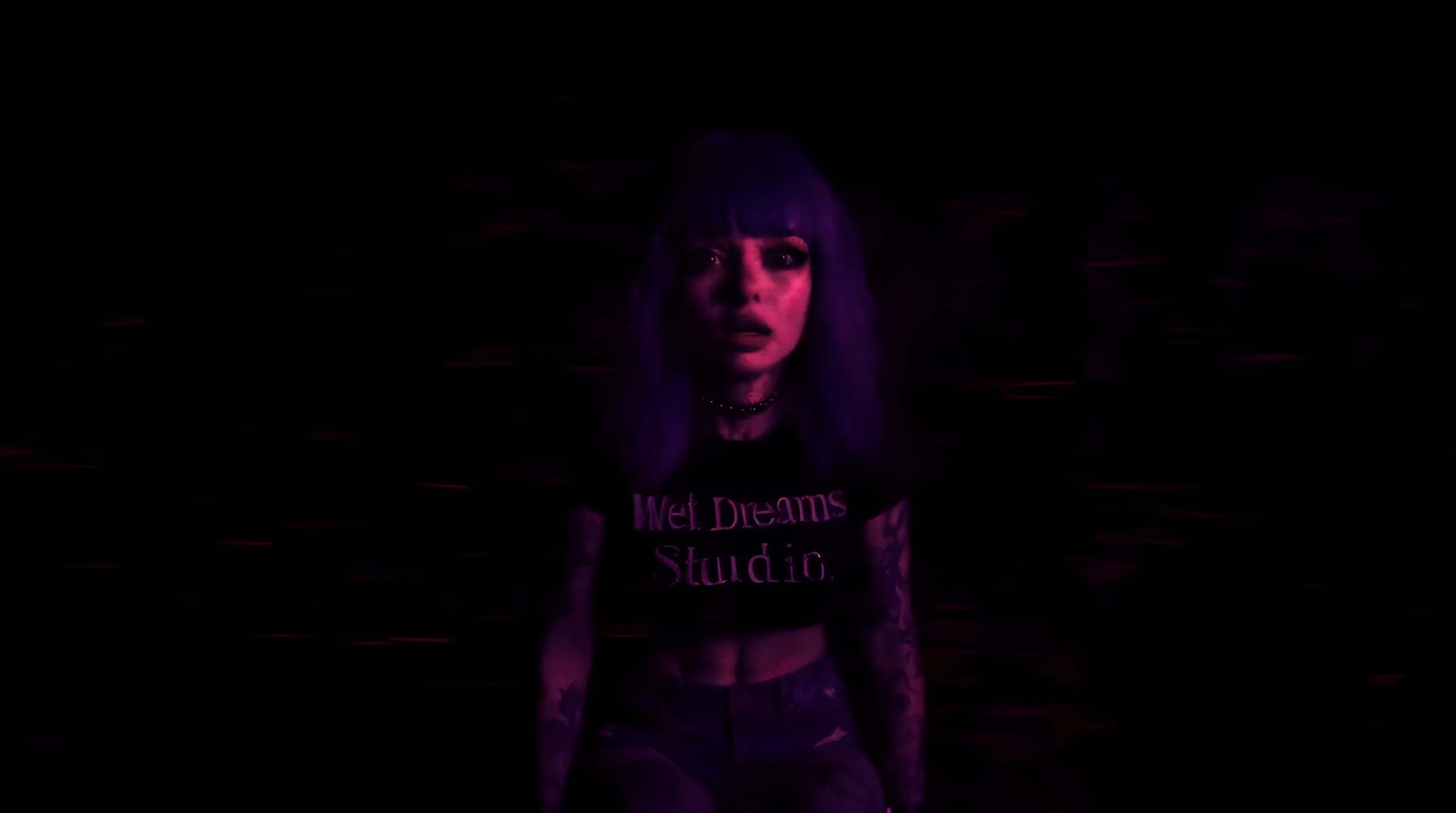 A woman with purple hair and tattoos stands in a dark setting illuminated by pinkish-purple lighting, wearing a black T-shirt with the words 'Wet Dreams Studio' in pink.