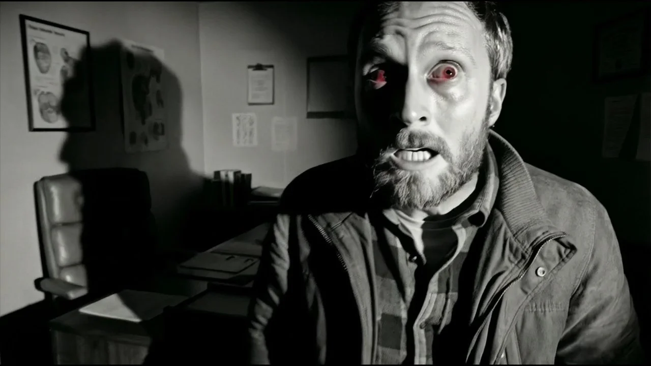 A man with a beard makes a frightened face in a dimly lit room, with an eerie shadow on the wall behind him.