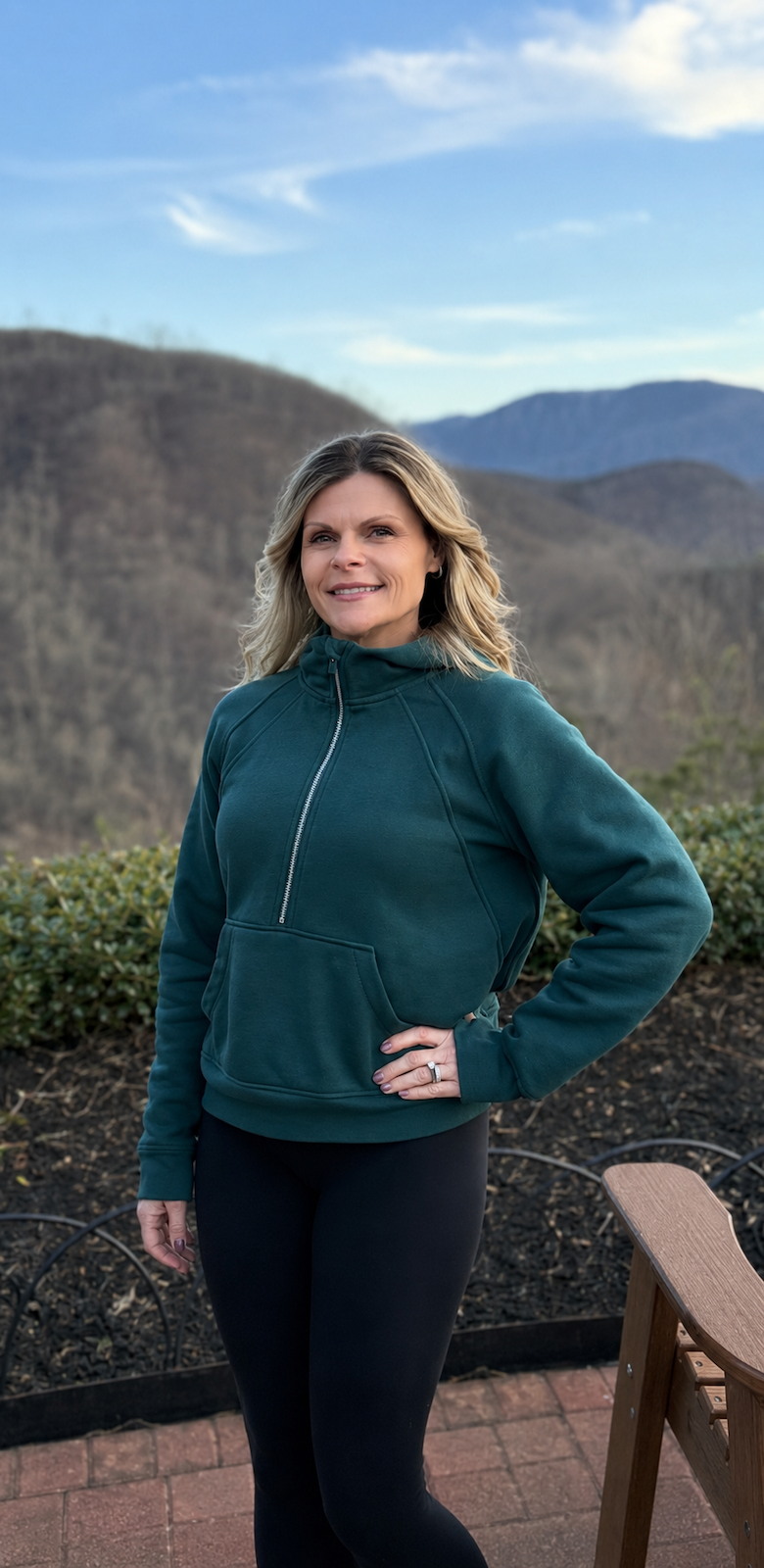 A woman standing outdoors on a patio with mountains and a blue sky in the background. She is wearing a teal zip-up jacket and black leggings, with her right hand on her hip, smiling slightly.