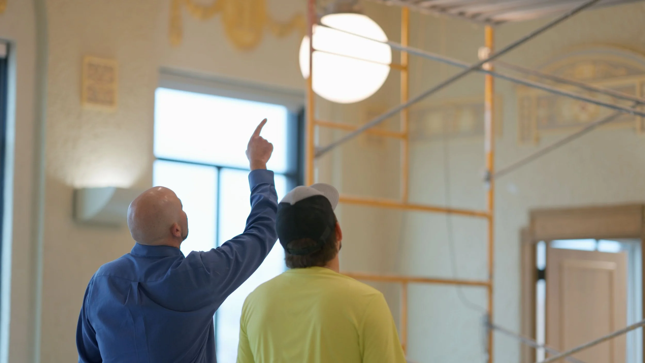 Two men are inside a building, one of them is pointing at something on the ceiling or above.