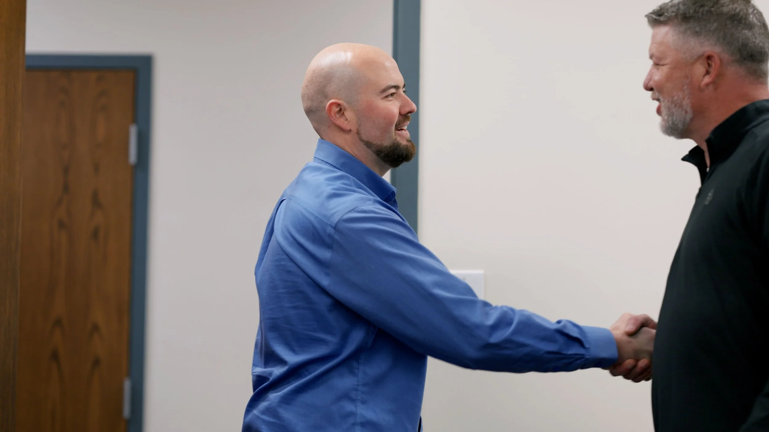 Two men are shaking hands and smiling in an indoor setting, one in a blue shirt and the other in a black shirt.