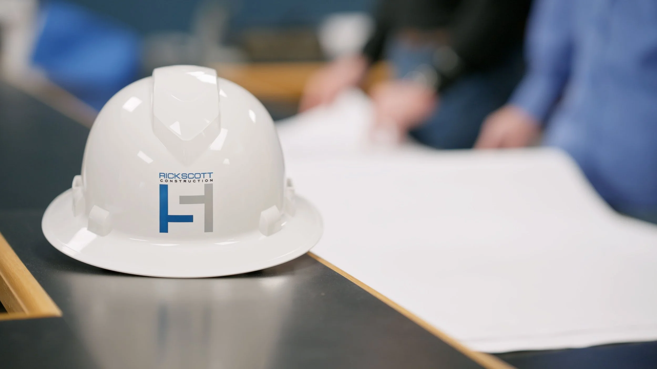 Construction helmet on a table with the logo 'Rick Scott Construction', with people and papers blurred in the background.