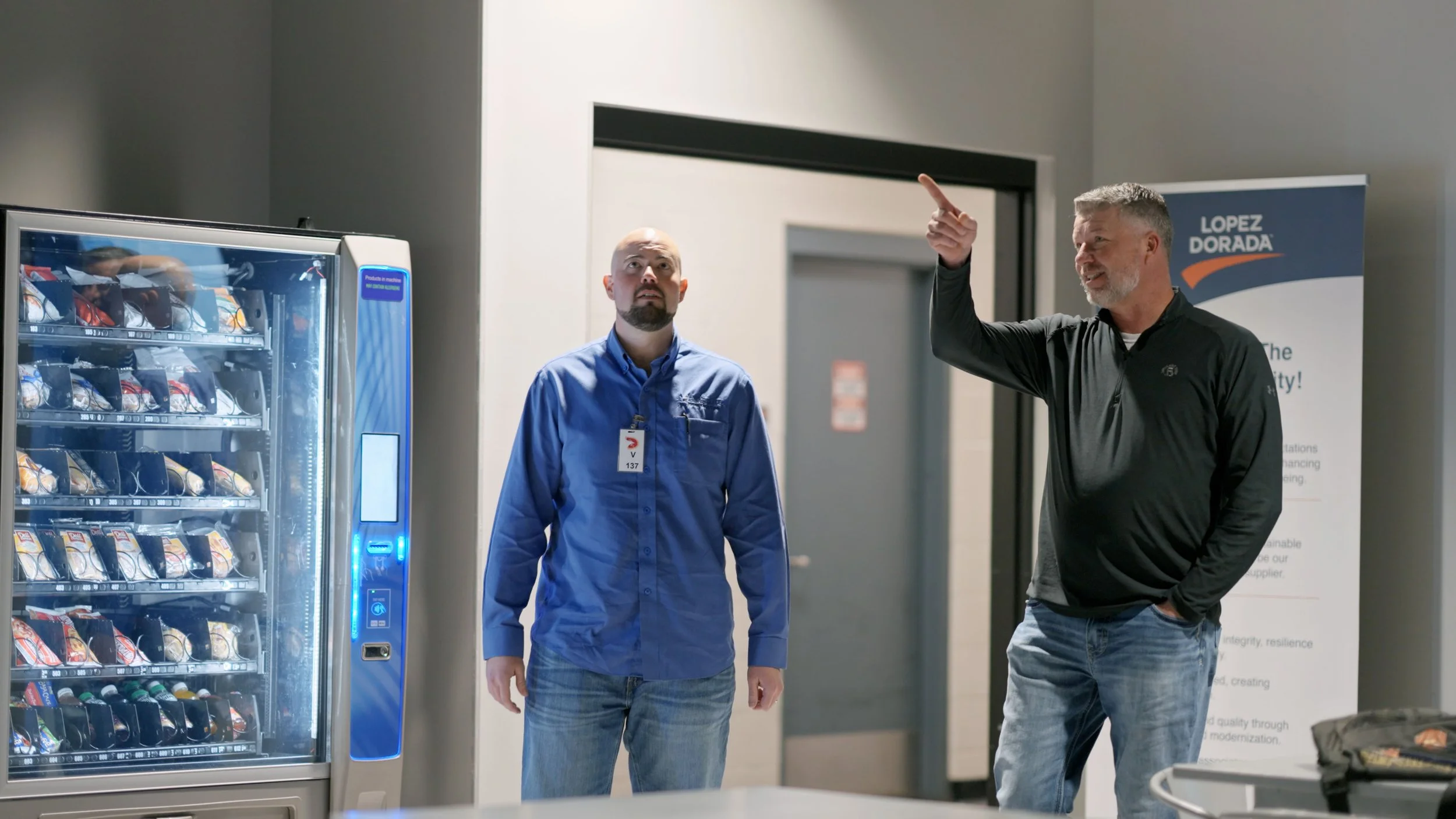 Two men standing near a vending machine in a modern office or public space, one gesturing with his hand and speaking, the other looking on. There is a banner in the background with the words 'Lopez Dorada' and an orange checkmark.