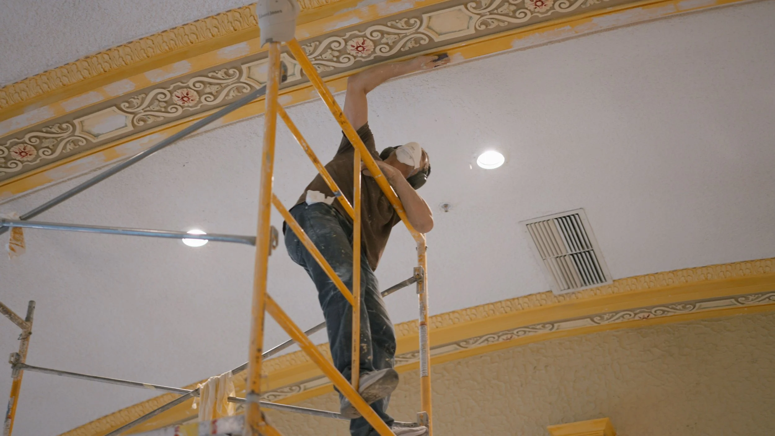 A worker standing on a yellow scaffolding, painting the crown molding on a ceiling with a paint roller. The ceiling has recessed lighting and an air vent.