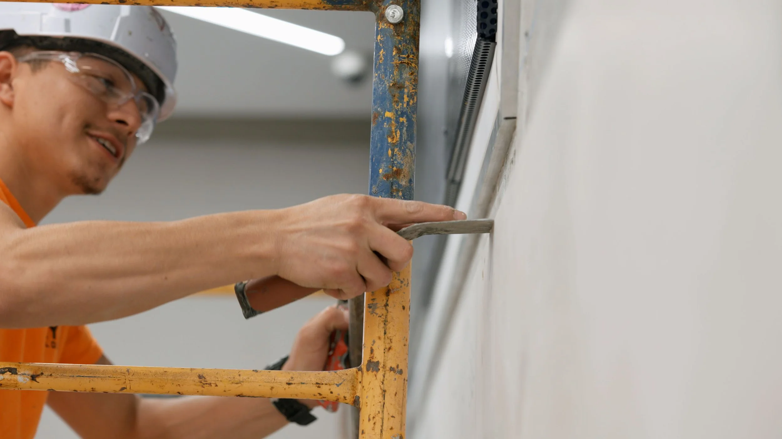 A man wearing safety goggles and an orange shirt is working on a wall with a paint scraper.