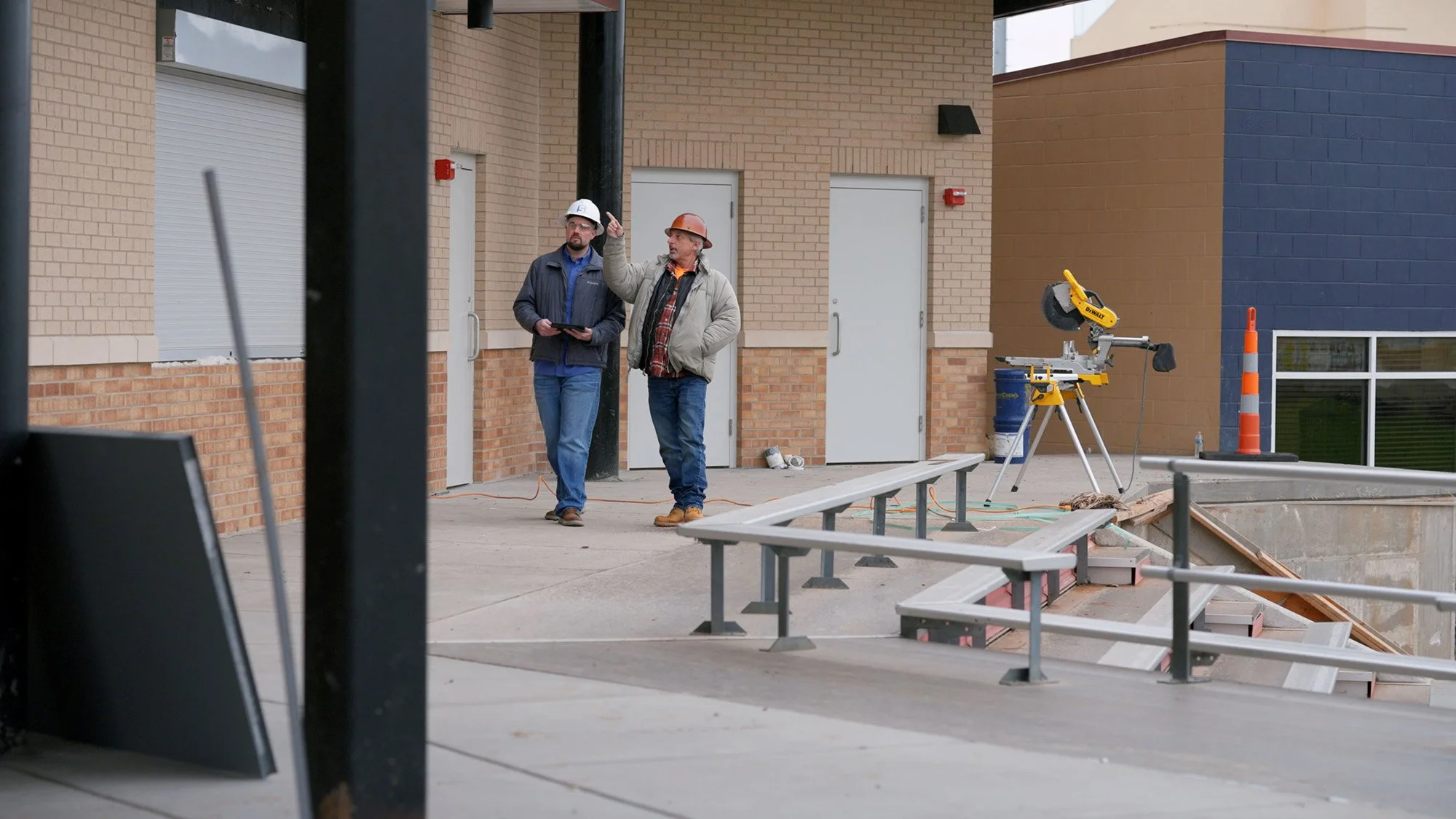 Two construction workers wearing safety helmets, one in orange and the other in white, are standing outside a building discussing something. Construction tools and equipment, including a miter saw, are visible around them, and there's a partially built structure with outdoor railings and a hole in the ground.