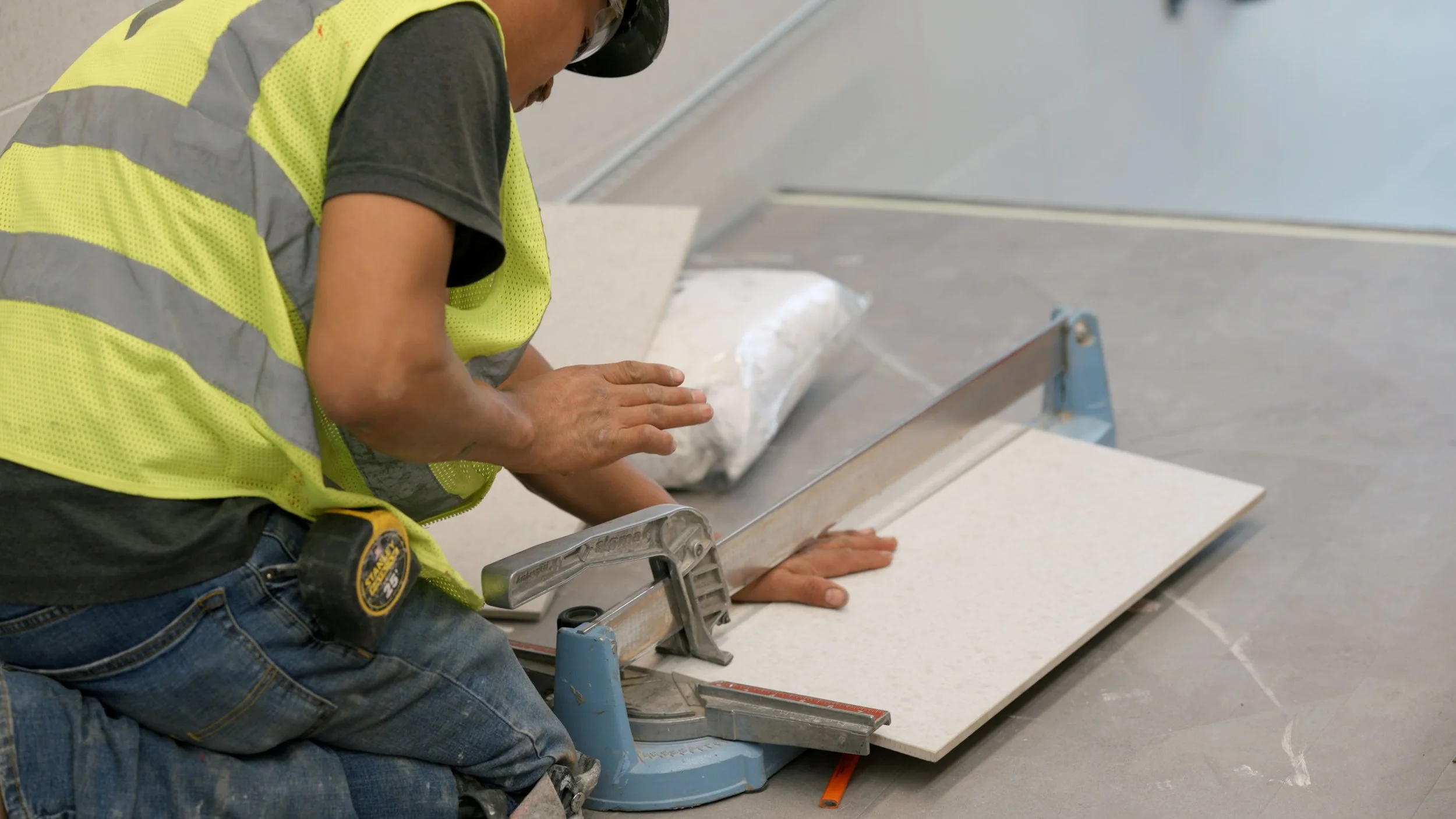 A construction worker wearing a yellow safety vest and a black hard hat is installing a large tile or countertop using a tile saw. The worker is kneeling on the floor, pressing down on the material while guiding it through the saw.