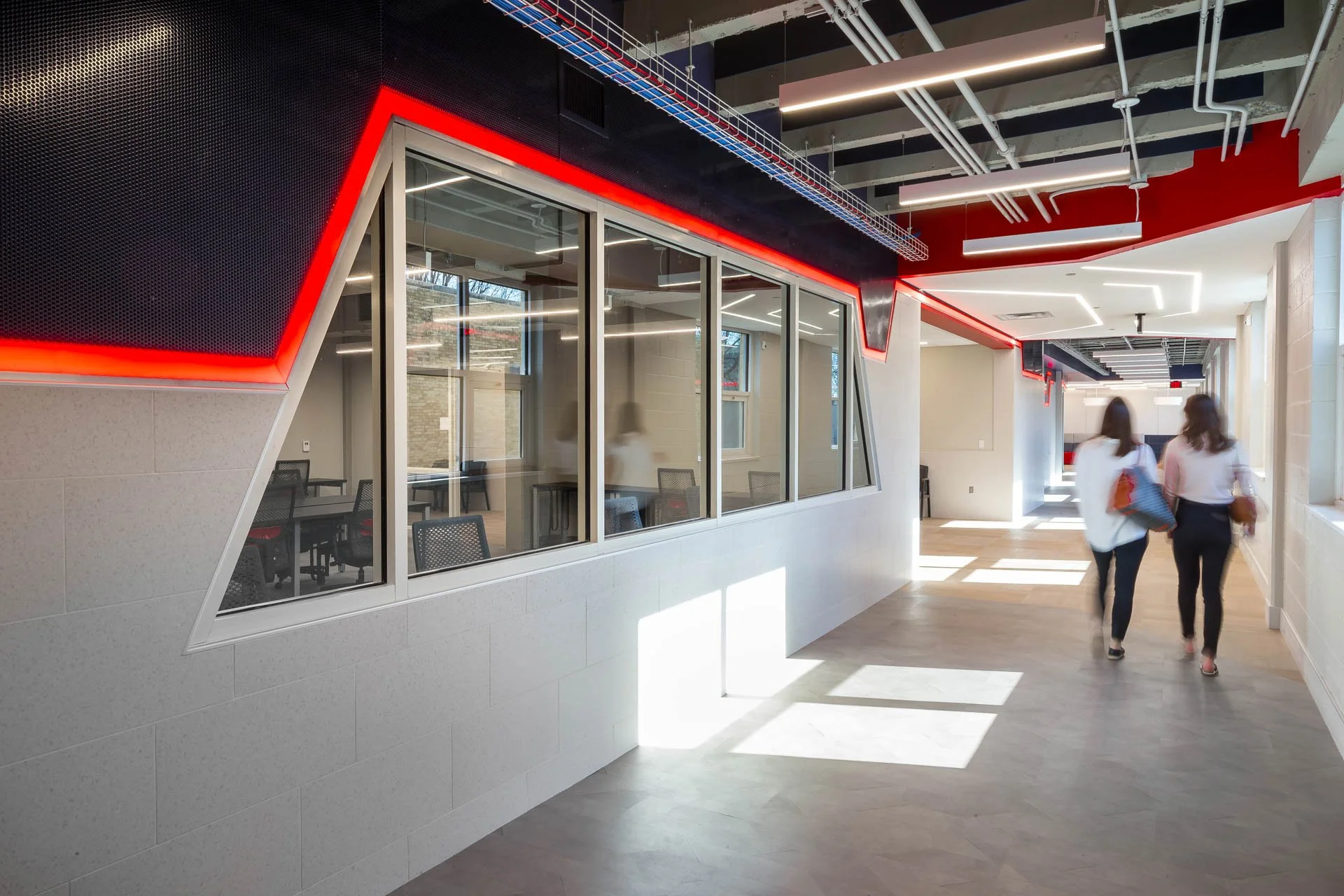 Modern office hallway with geometric ceiling lighting and exposed piping. Two women walk together, carrying bags, near glass-walled conference rooms on the left.
