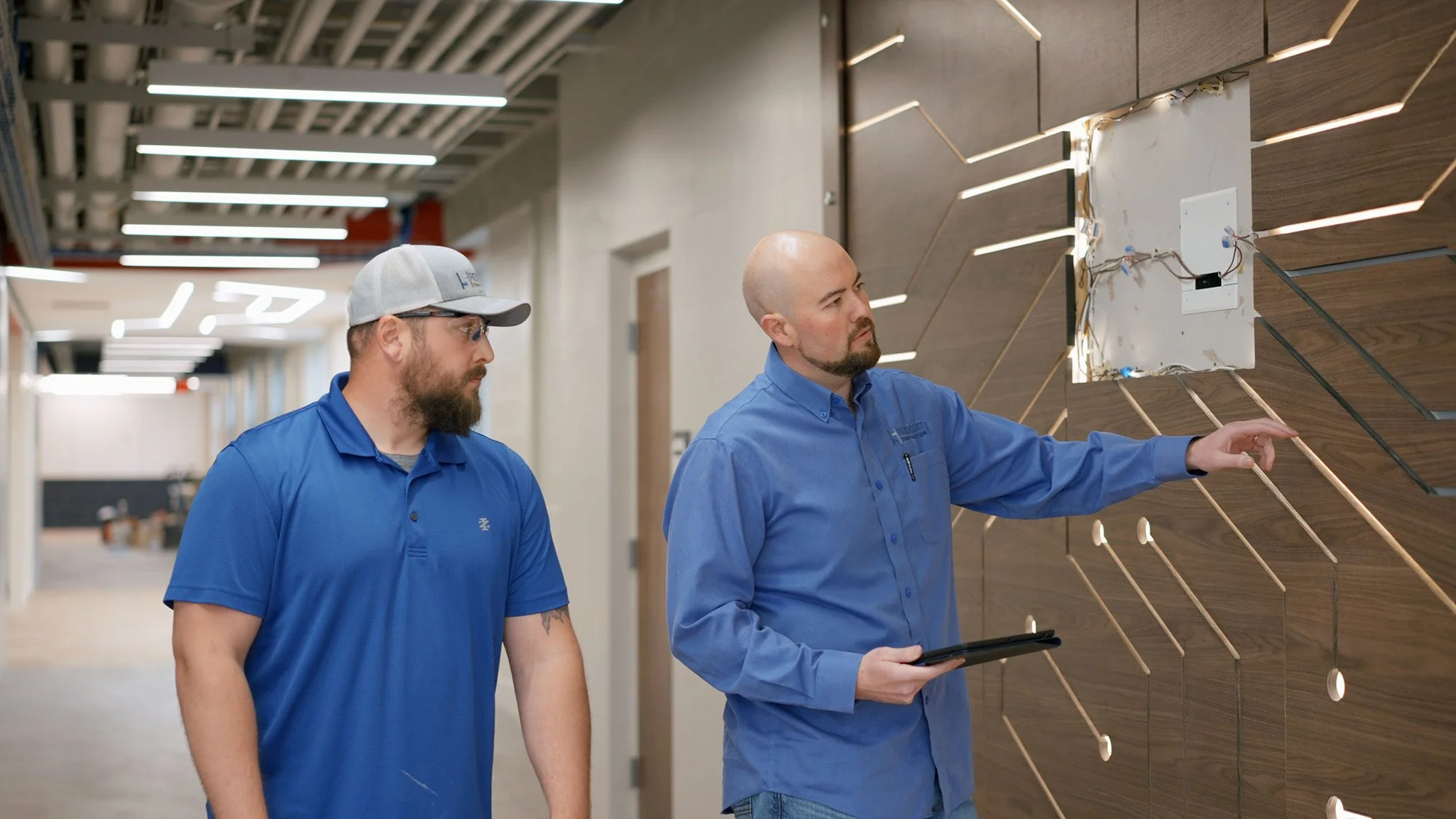 Two men in blue shirts standing in front of a wooden wall with illuminated lines, one holding a tablet and pointing at the wall, the other listening.