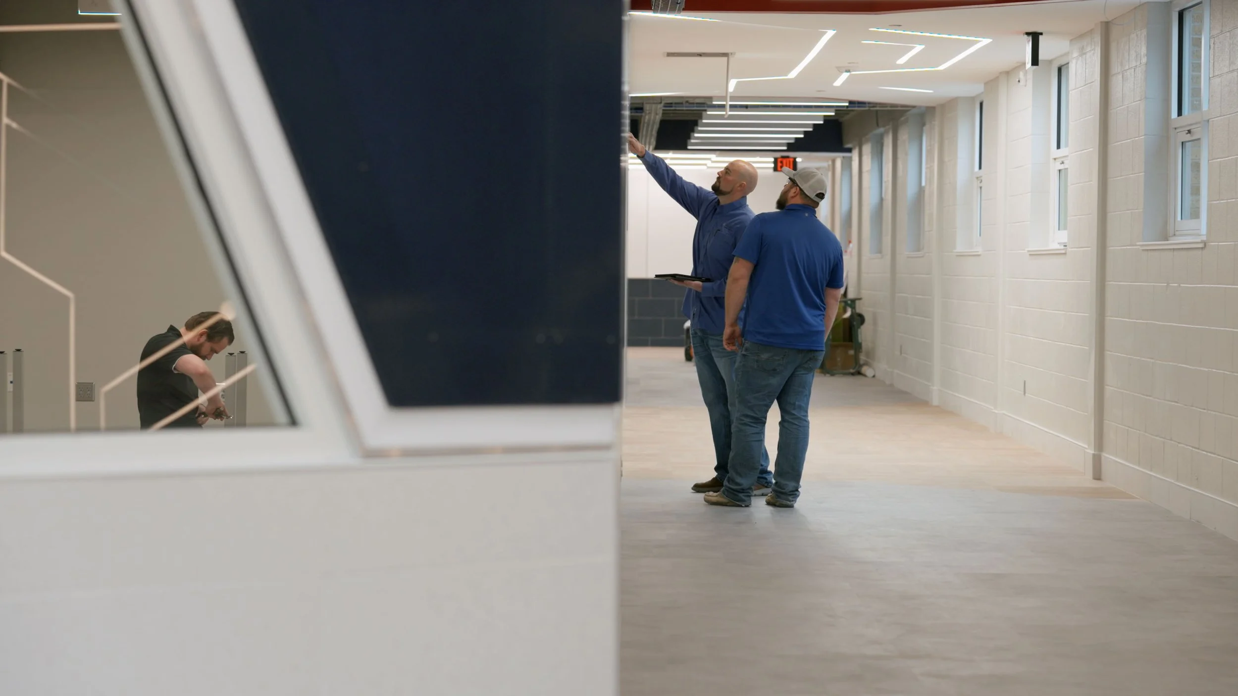Two men in blue shirts and jeans are standing in a hallway, one pointing at the ceiling, while another man in a gray cap looks on; a person in black is visible through a glass window or door on the left side.