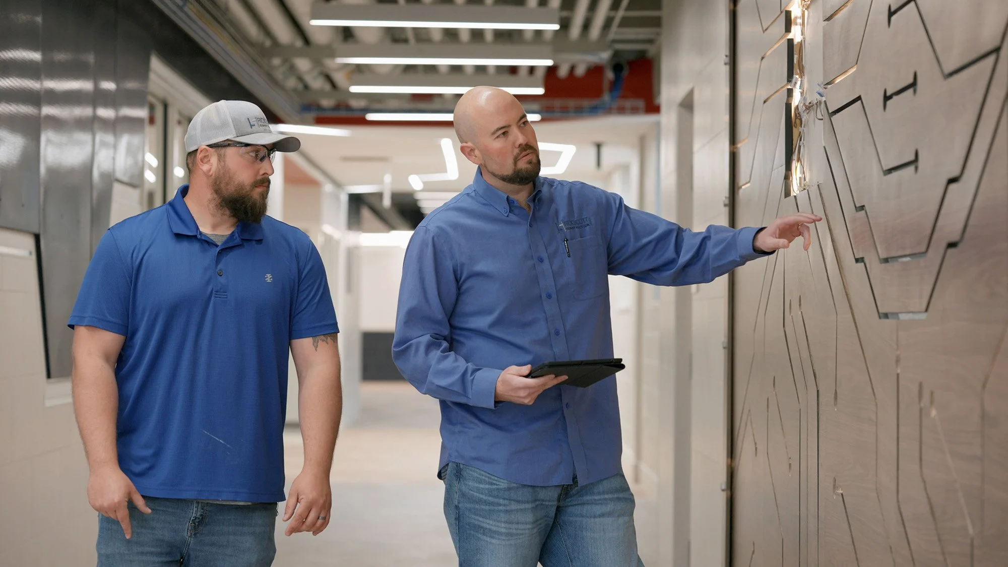 Two men working in an industrial or corporate setting, inspecting a wall with geometric designs and wiring. One man is pointing at the wall, holding a tablet, while the other listens attentively.