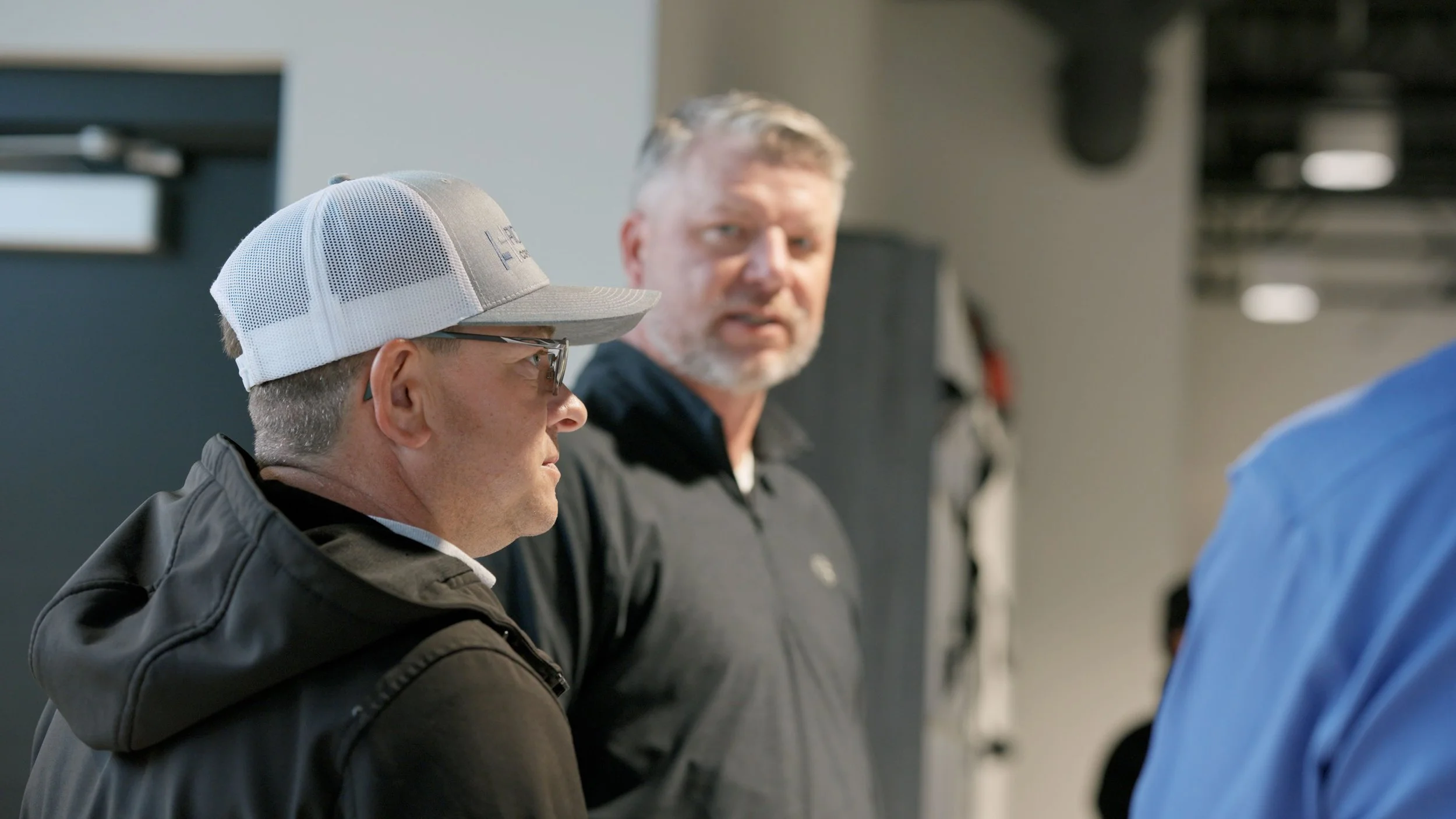 Three men engaged in a conversation indoors, with one man facing sideways wearing a white baseball cap with mesh back, another man in the background has gray hair and a beard, and the third man partly visible in the foreground with a blue shirt.