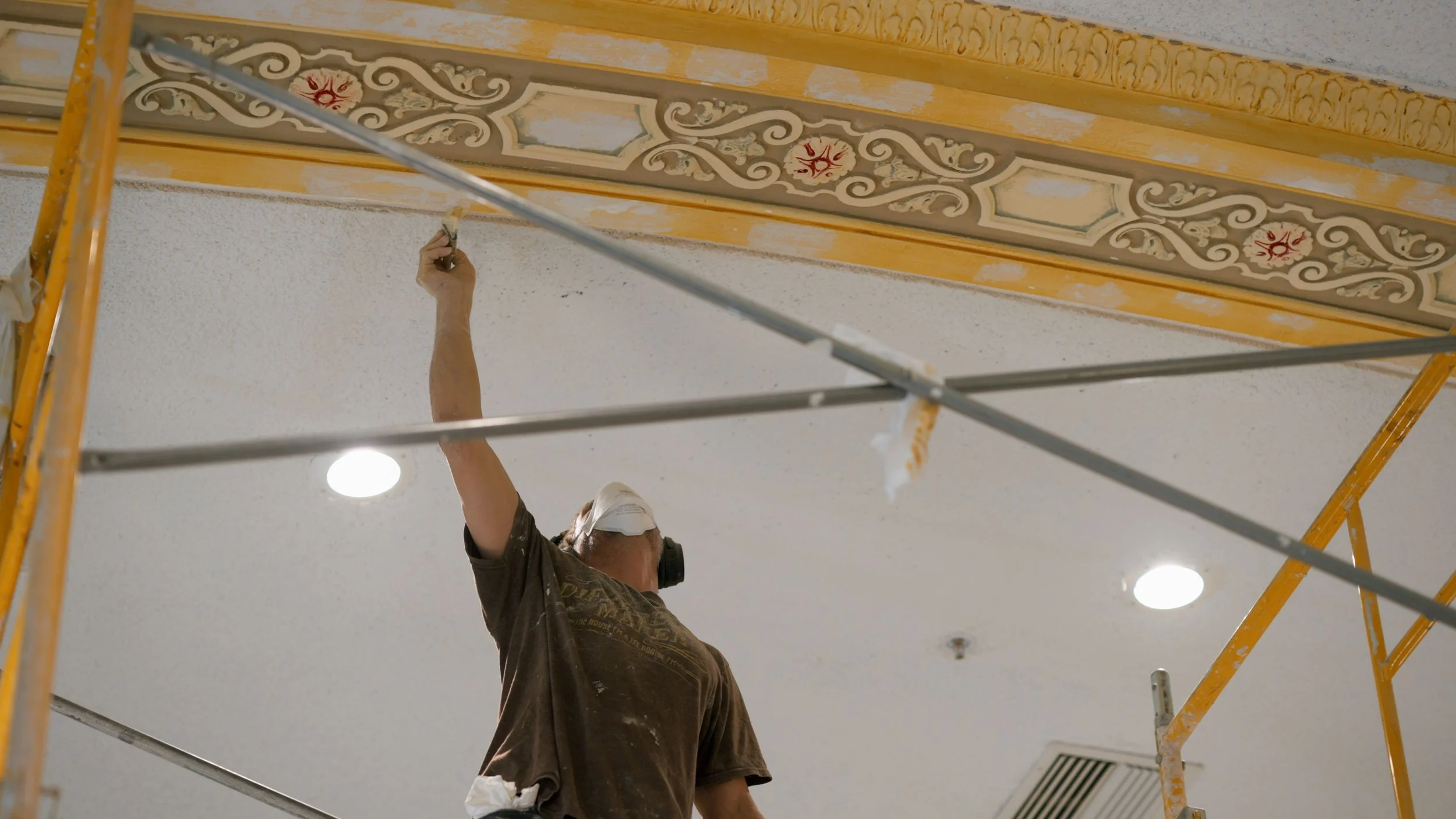 A worker painting the ceiling decorative trim on a scaffolding inside a building, wearing a mask and protective gear.