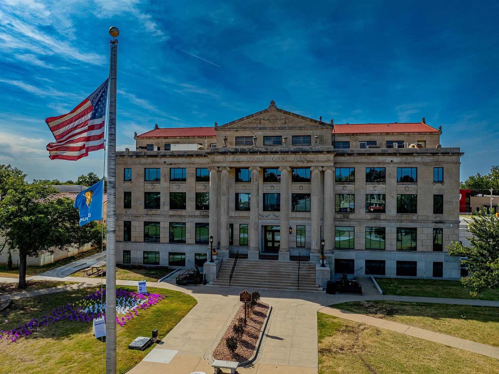 The Jackson County Courthouse, a historic building with stone architecture, tall columns at the entrance, and red tiled roof, is visible under a blue sky with clouds. Flags of the United States and a blue flag are flying in front.