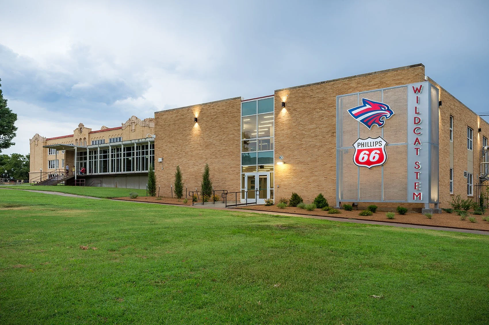 A modern brick building with a sign displaying the Wildcat Stadium logo and the word 'Wildcats Stadium' on a vertical red sign. The building has large windows, greenery, and a grassy foreground under a cloudy sky.