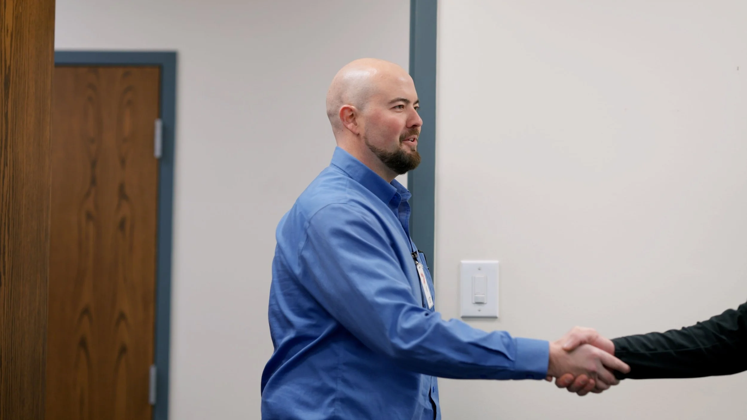 A man with a shaved head and beard wearing a blue shirt, shaking hands with another person in a professional setting.
