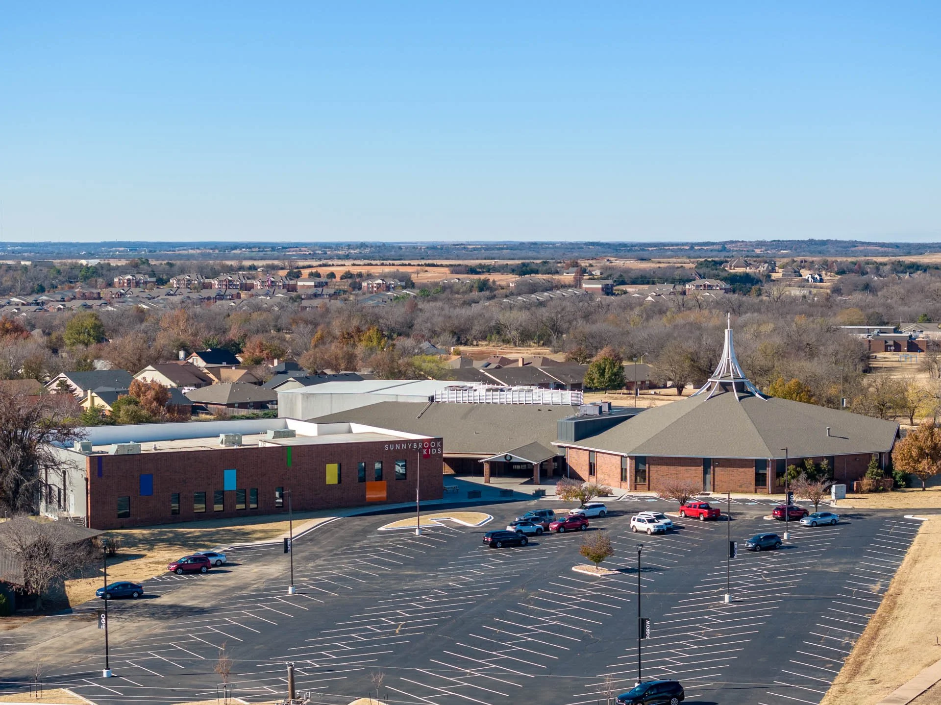 Wide view of a mostly empty parking lot with a few parked cars, a building with a sign reading 'Sunnybrook', and a large building with a distinctive pointed roof in the foreground. Beyond the parking lot, suburban houses and trees stretch to the horizon under a clear blue sky.