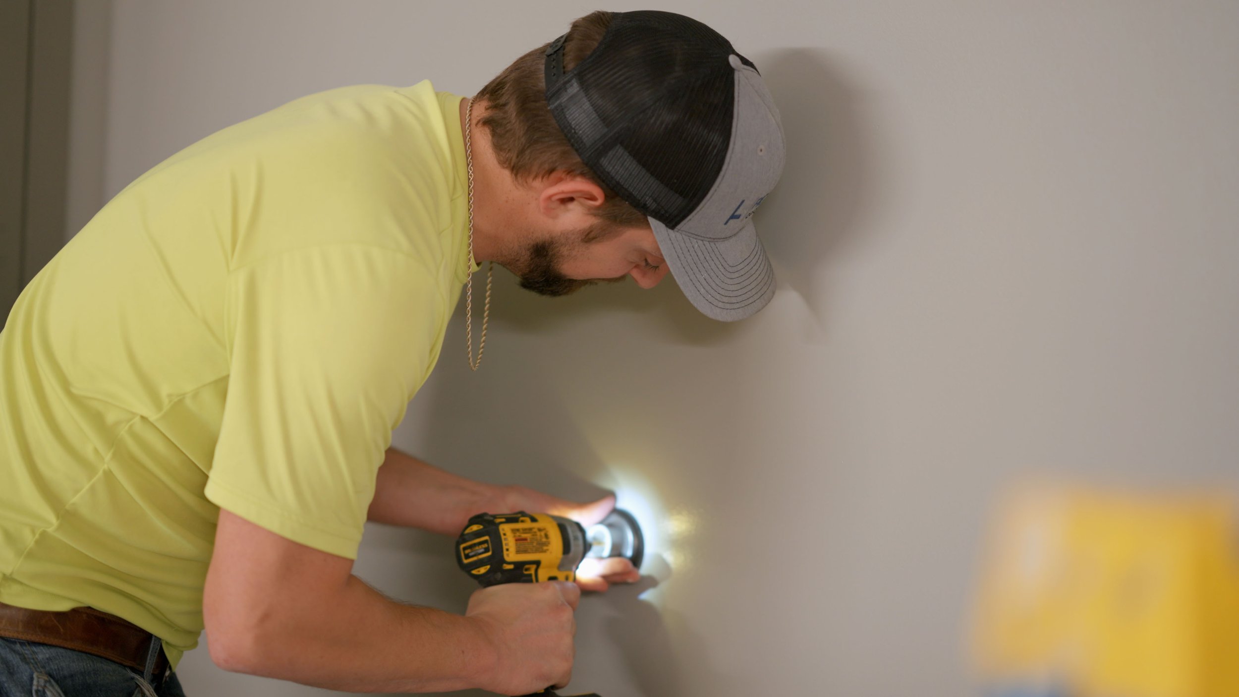A man wearing a gray and black trucker hat, a yellow t-shirt, and a silver chain is using a yellow and black cordless drill on a wall. The man appears to be installing or fixing something in the wall, and there is a flashlight providing illumination.
