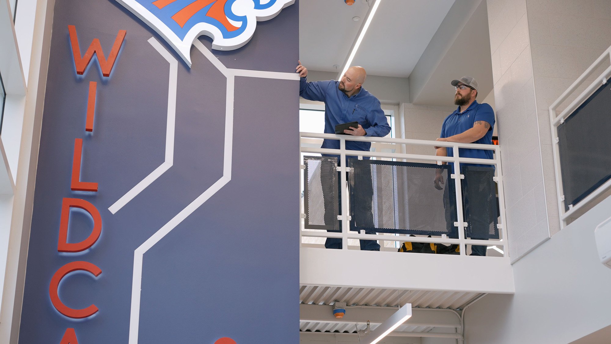 Two men in blue shirts are standing on a balcony-like structure inside a building, looking at a large wall logo. One man is touching the logo with his hand, and the other is observing. The background features large windows and modern interior design.