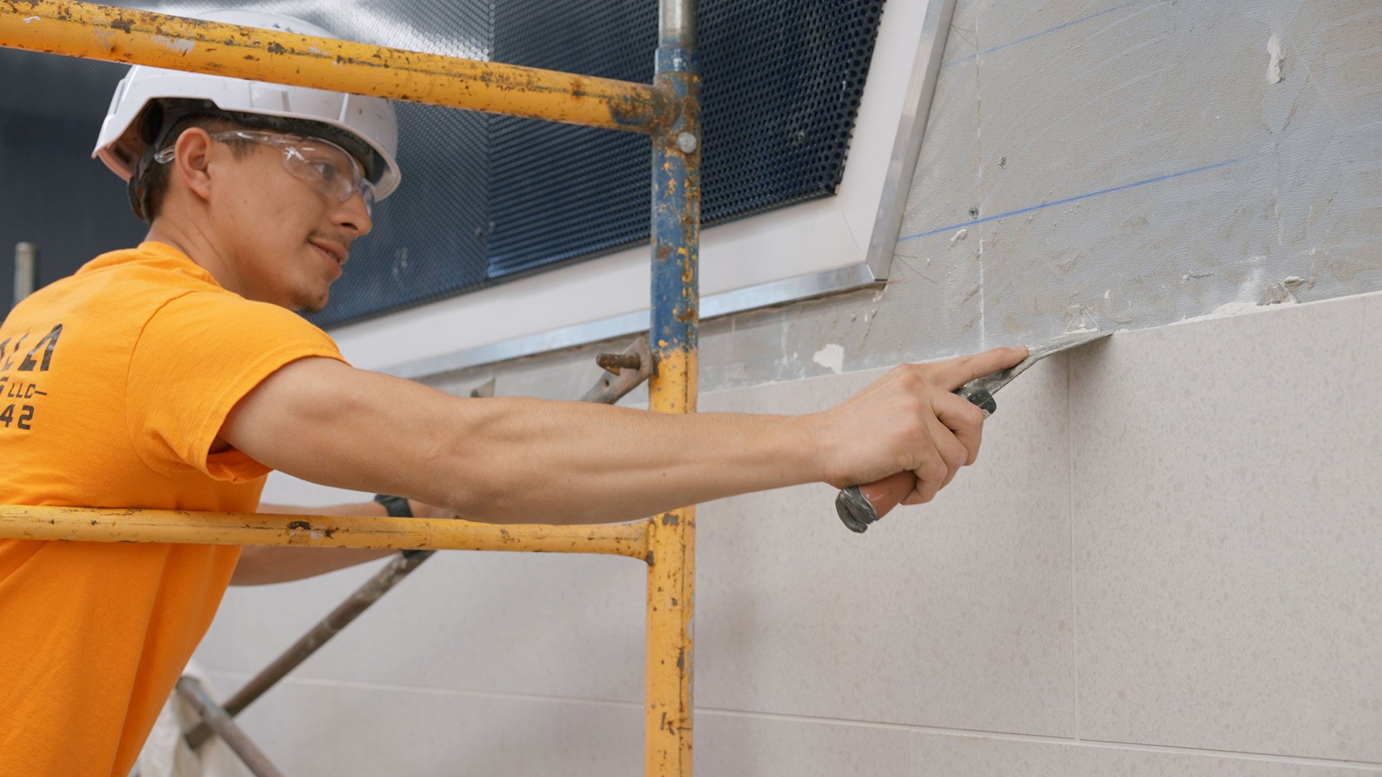 A construction worker wearing safety glasses and a white helmet uses a trowel to apply a finishing coat to a wall while standing on a yellow scaffold.